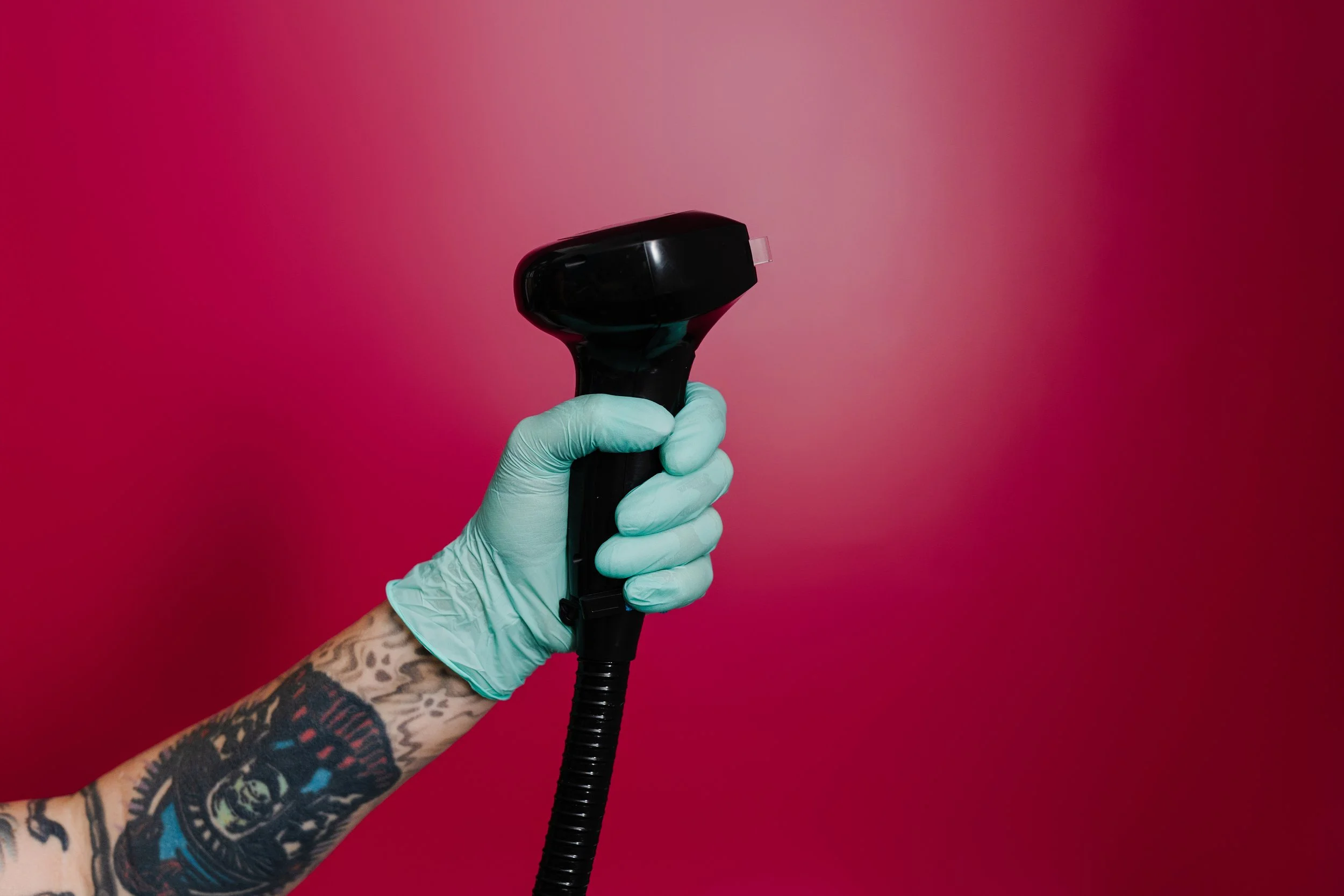 A tattooed person wearing a light blue glove holding a black handheld device against a pink gradient background.