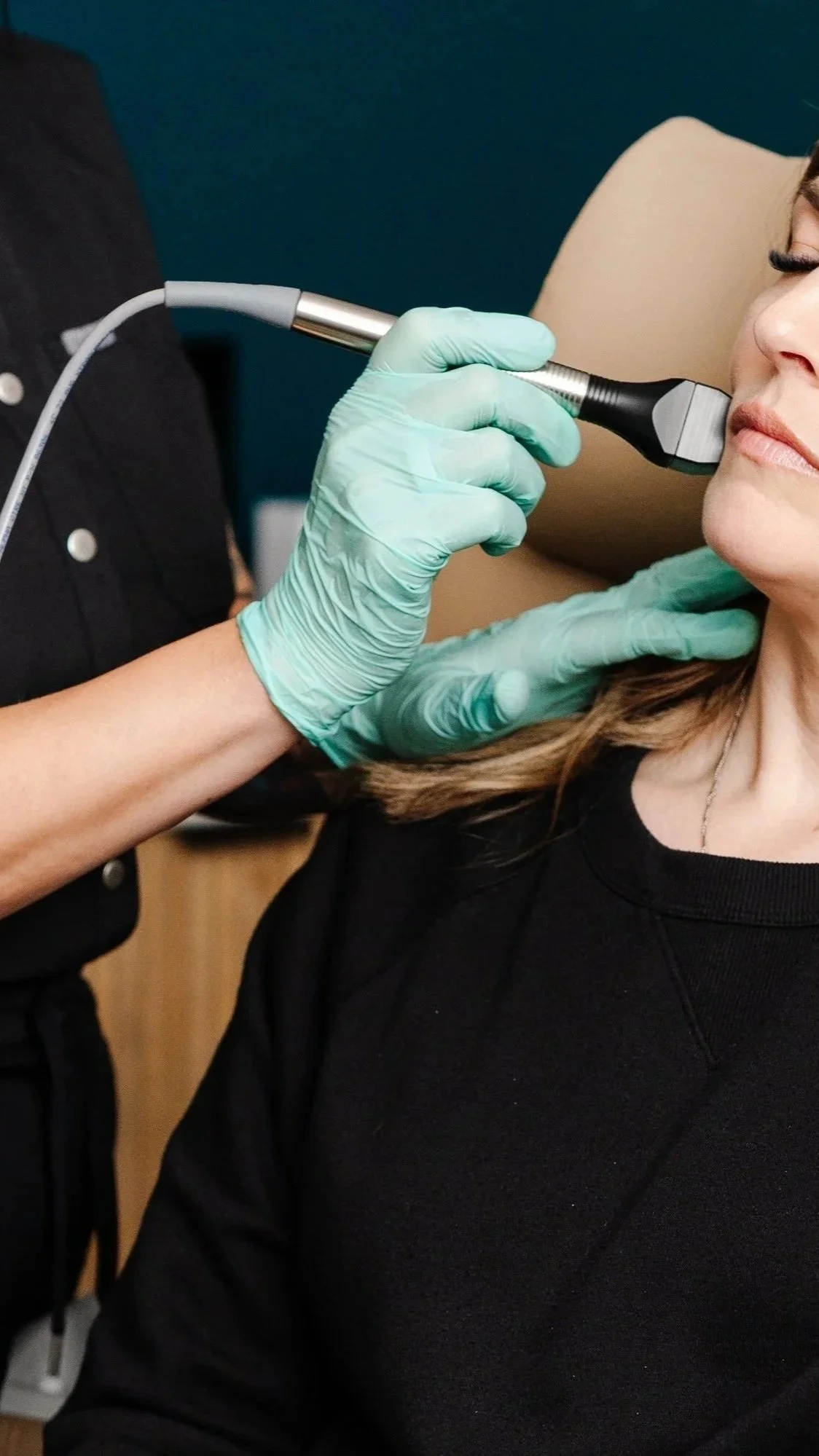 A woman receiving a facial treatment with a handheld device from a professional wearing teal gloves.