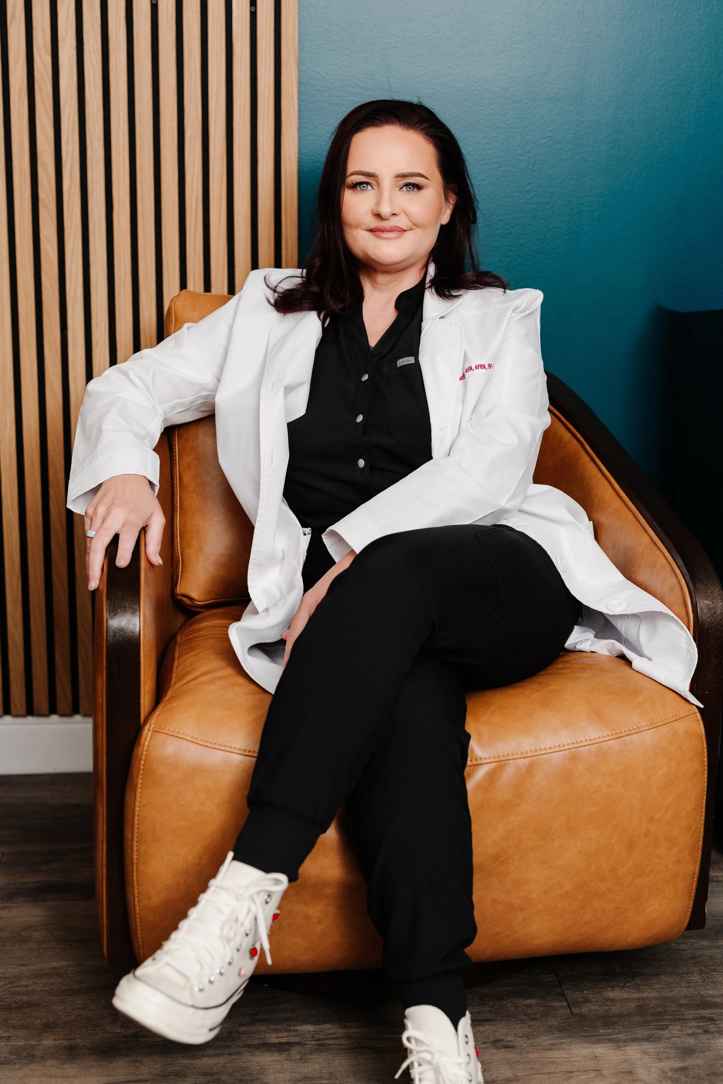 A woman with dark hair and blue eyes sitting on a tan leather chair, wearing a white coat over a black shirt, black pants, and white sneakers, in a room with a teal wall and wooden paneling.
