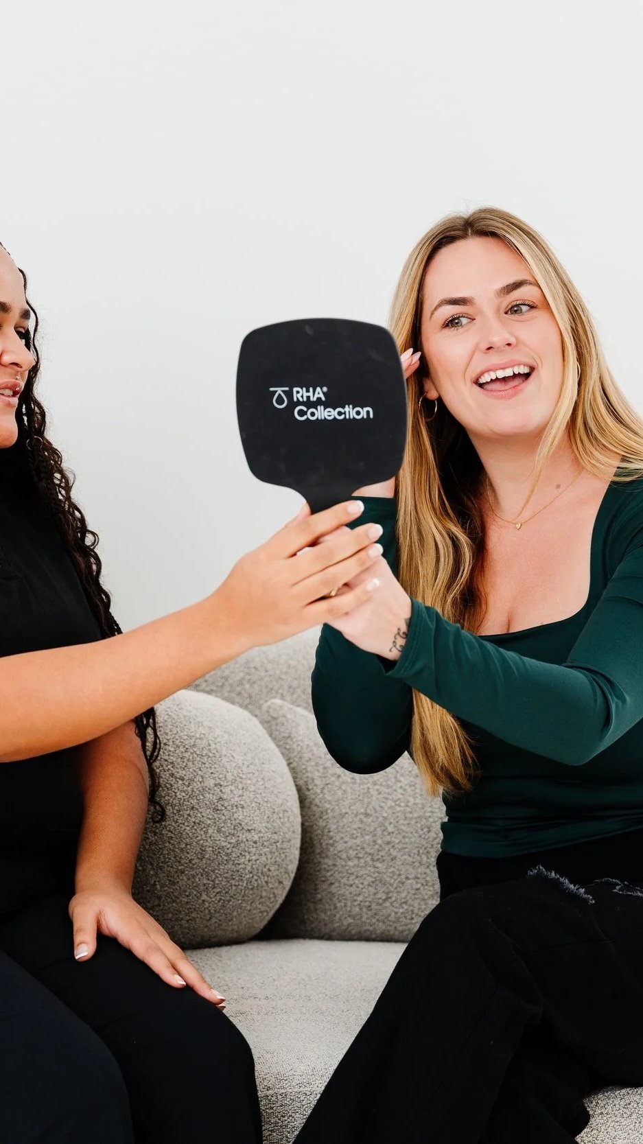 A woman with long blonde hair holding a mirror labeled 'RHA Collection,' sitting on a gray couch, smiling and talking to another woman with curly dark hair, wearing black.