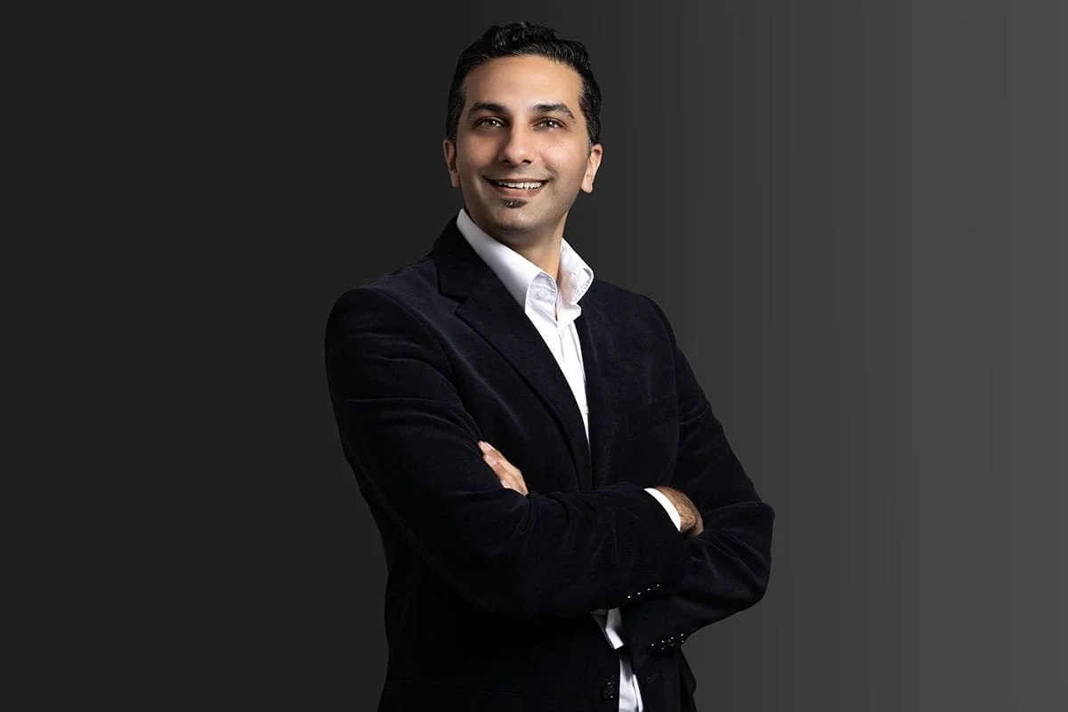 Professional man with dark hair and beard, wearing a white shirt and black blazer, smiling with arms crossed against a gradient dark background.