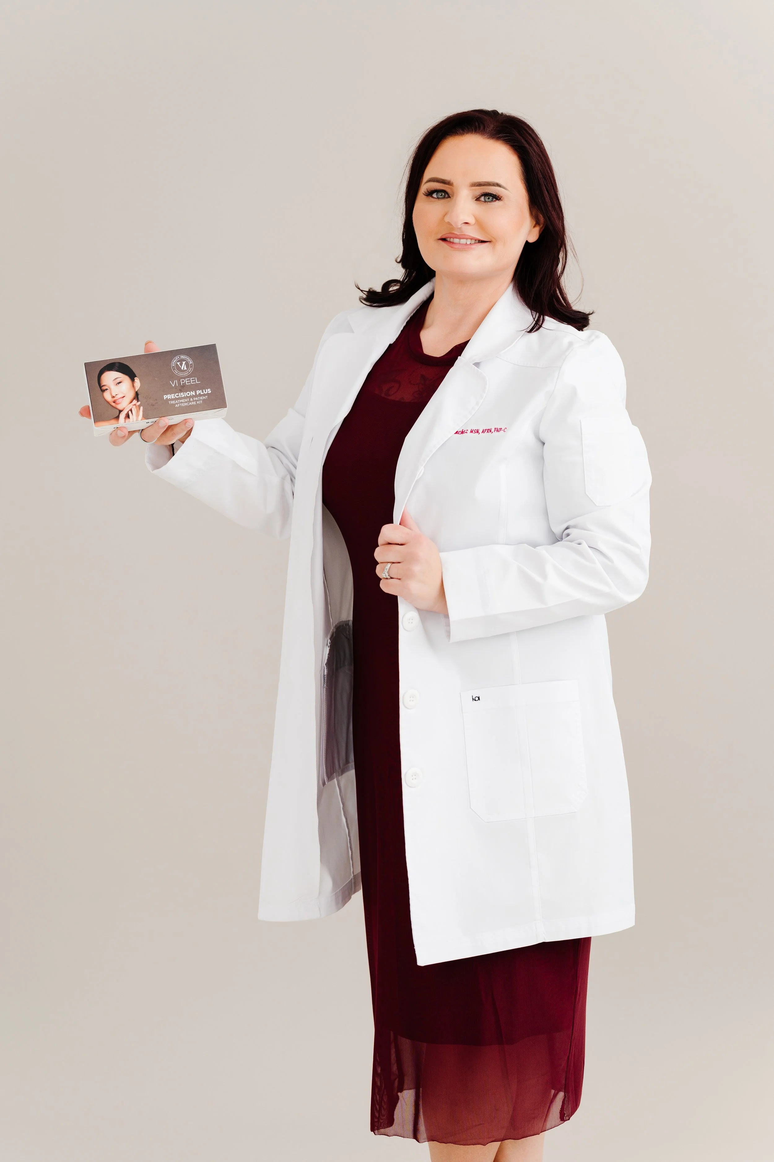 A woman with dark hair and light skin smiling, wearing a white medical coat and holding an informational card about a skincare treatment.
