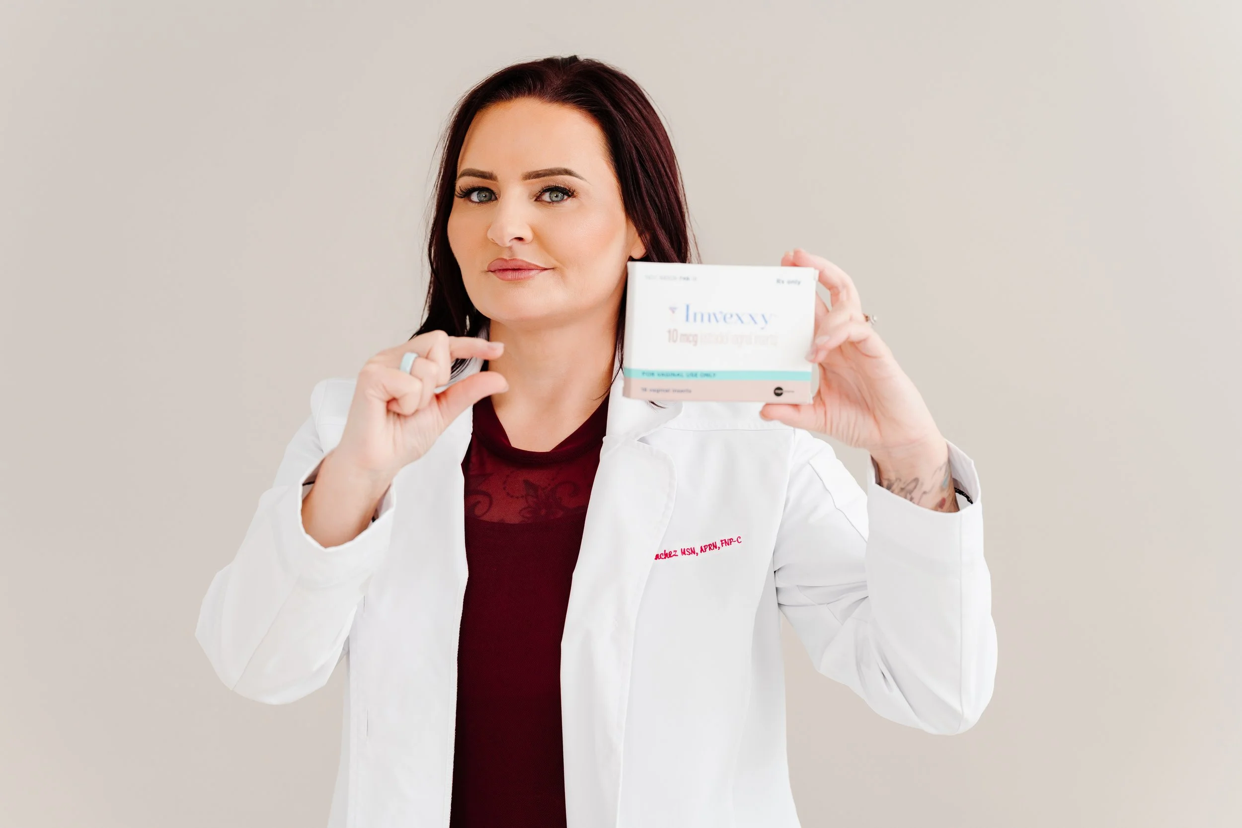 A woman with reddish-brown hair and light skin wearing a white medical coat holding a box of Invzzy medication, pointing at the box with her right index finger, against a plain beige background.
