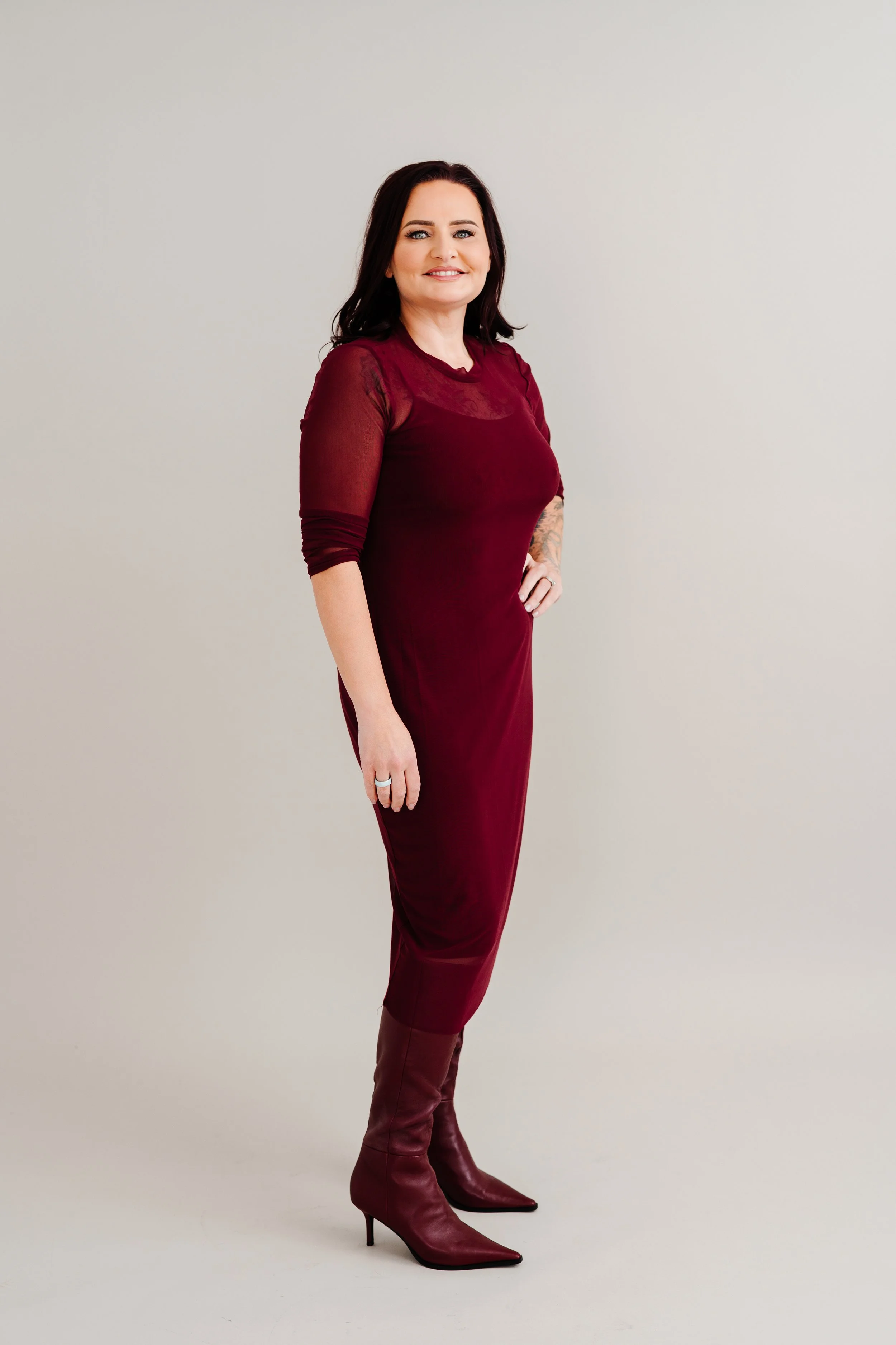 A woman dressed in a burgundy dress and matching high-heeled boots standing against a plain light gray background, smiling and looking at the camera.