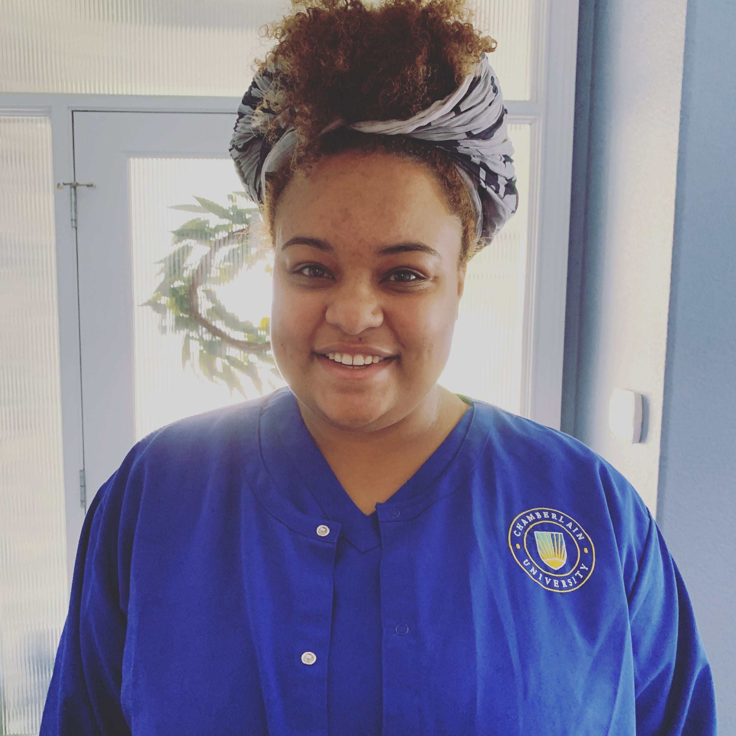 A woman with curly hair wrapped in a striped headscarf, wearing a blue shirt with an embroidered emblem from Chamberlain University, smiling near a window with plants outside.