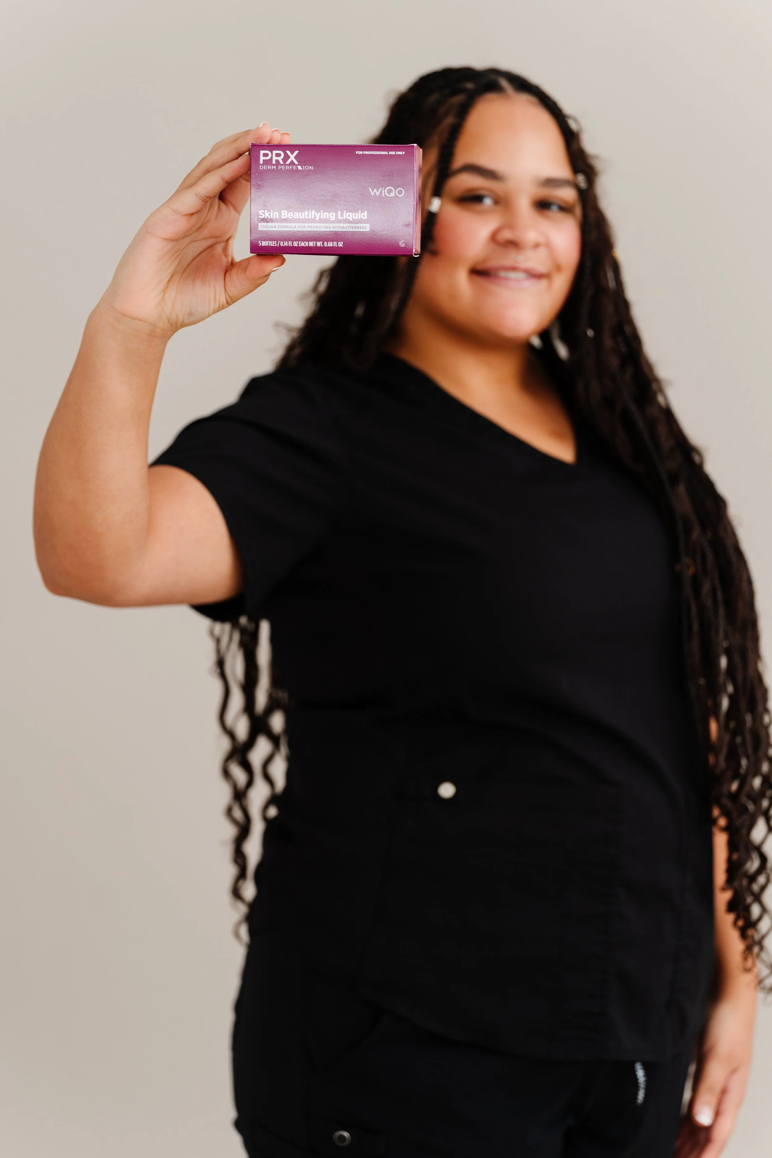 A woman with curly hair wearing a black shirt holds a pink box of skincare product labeled 'PRX Derm Perfexion Skin Beautifying Liquid' in front of a plain light-colored wall.