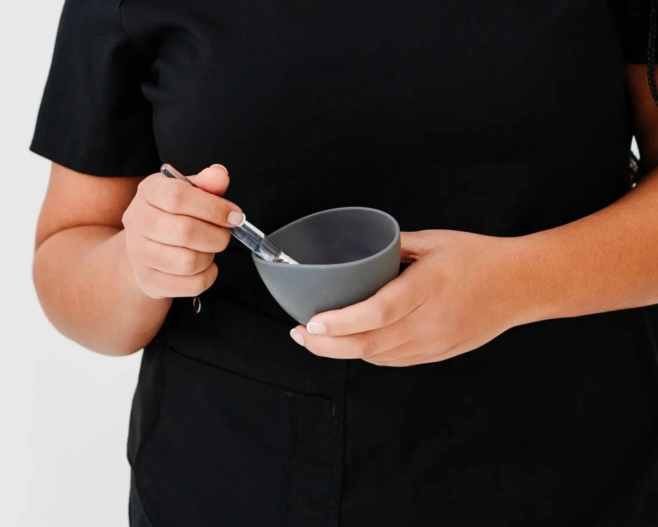A person wearing a black shirt is holding a gray bowl in one hand and a small spoon in the other, suggesting they are preparing to eat or mix something.