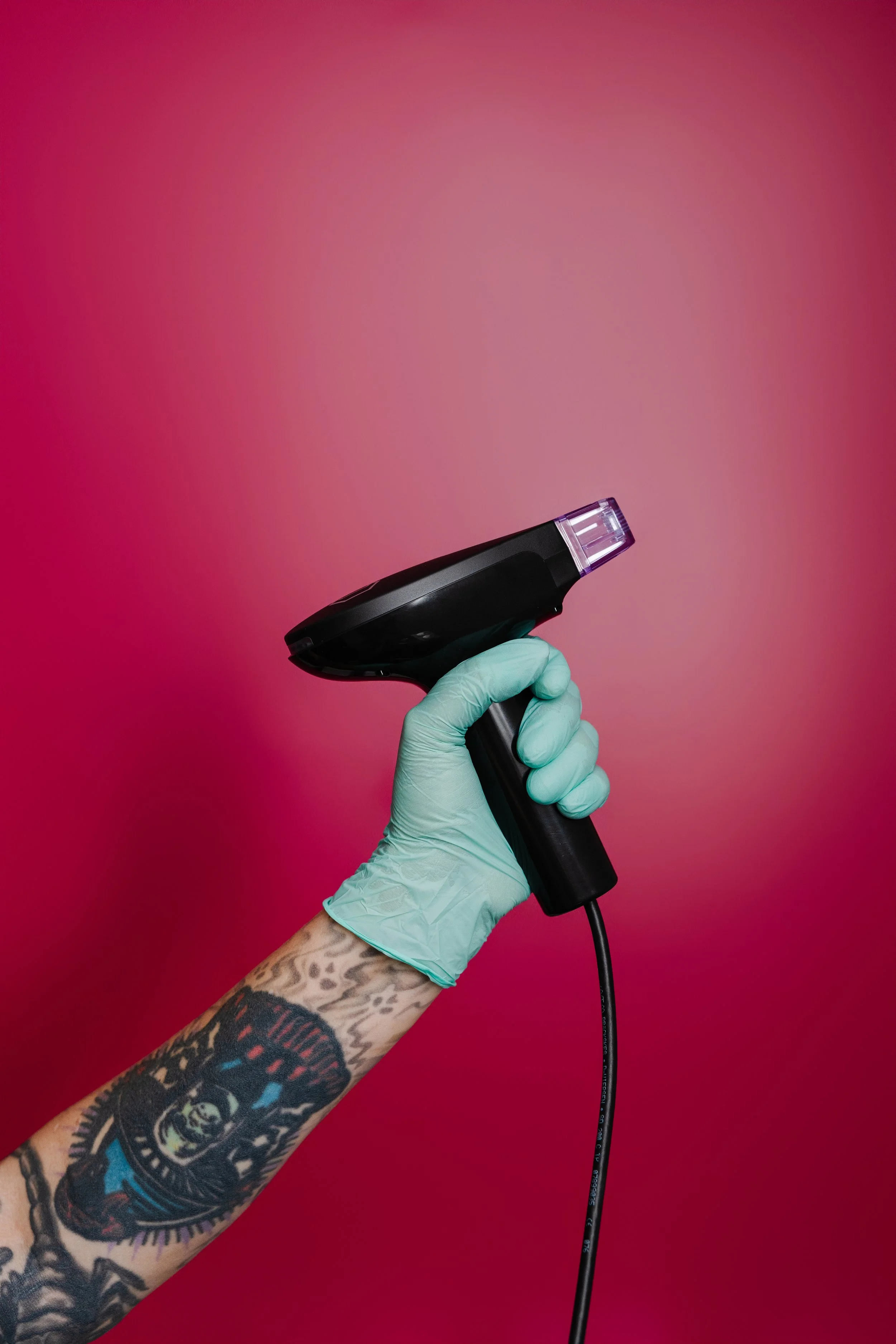 A hand wearing a green medical glove holding a black infrared thermometer against a pink background.
