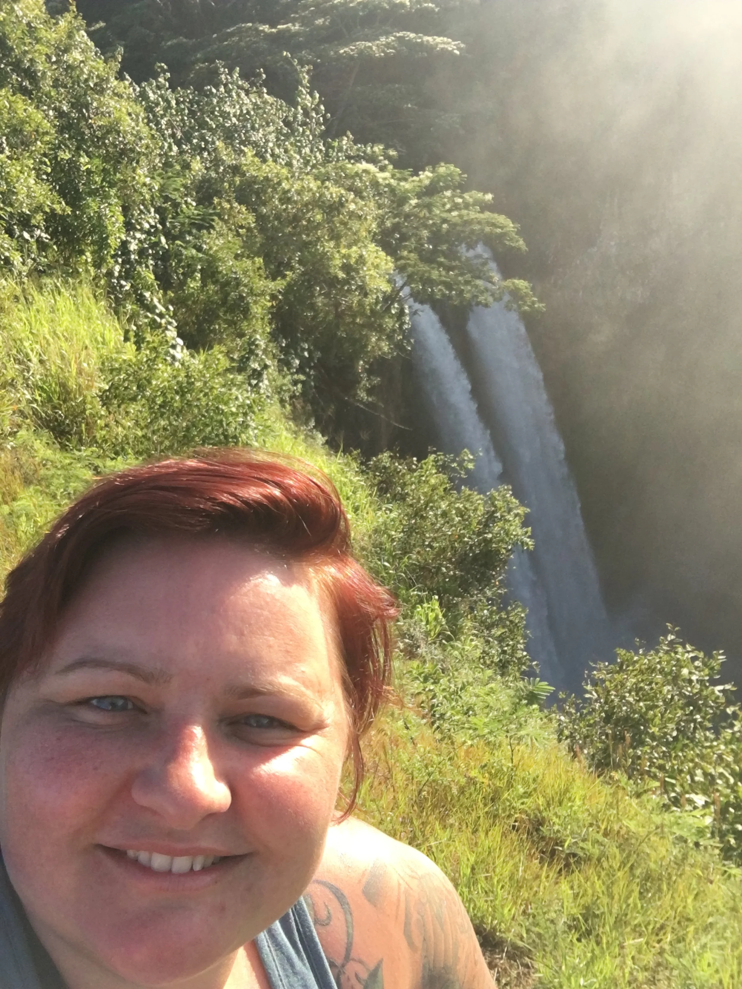 Person smiling with a view of a waterfall and surrounding greenery in the background.