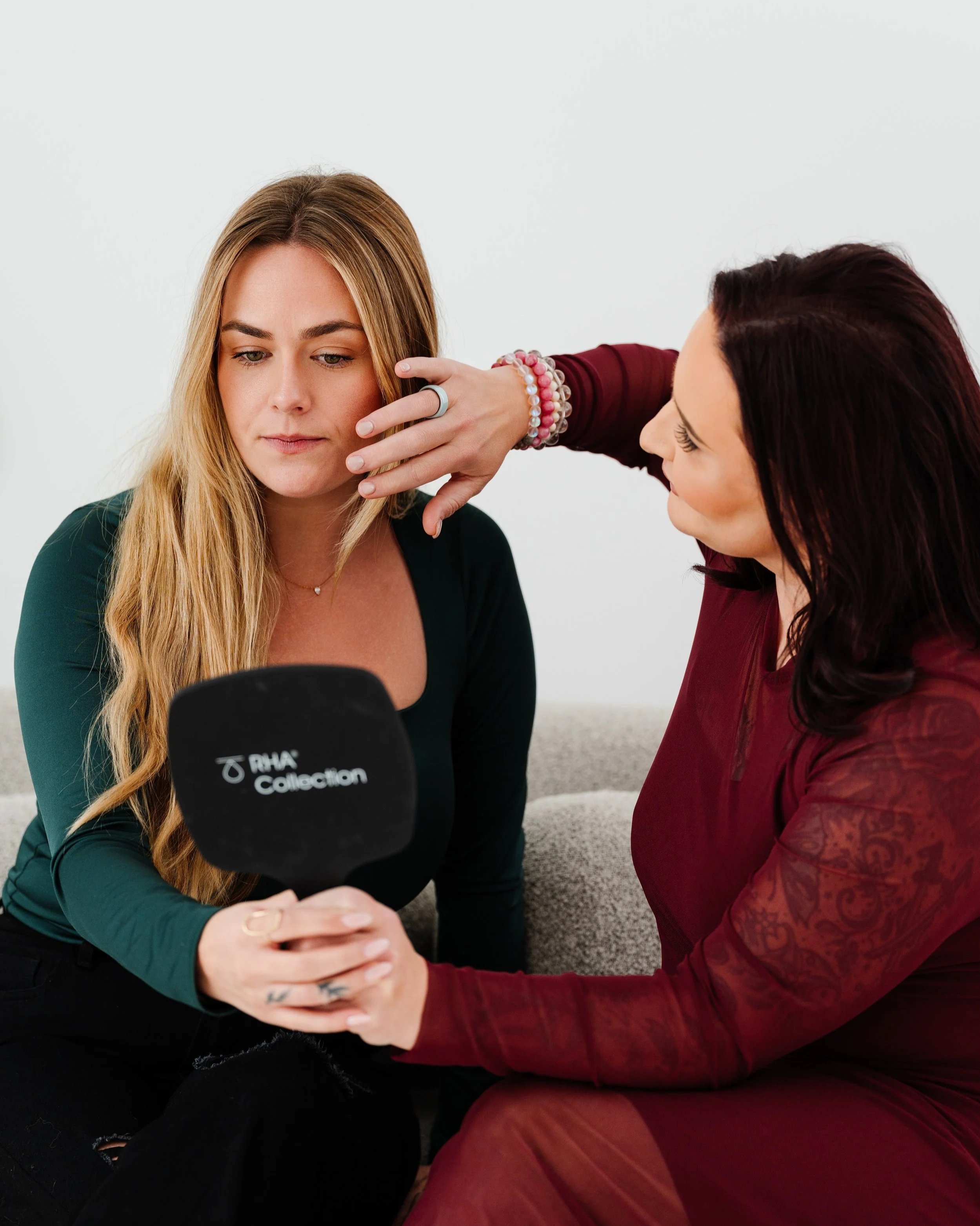Two women are sitting on the floor, closely looking at a smartphone together. One woman has long blond hair, and the other has dark red hair. The blond woman is wearing a dark green top, and the dark red-haired woman is wearing a maroon patterned dress. There is a black microphone with the logo of 'RVA Collection' in the foreground.