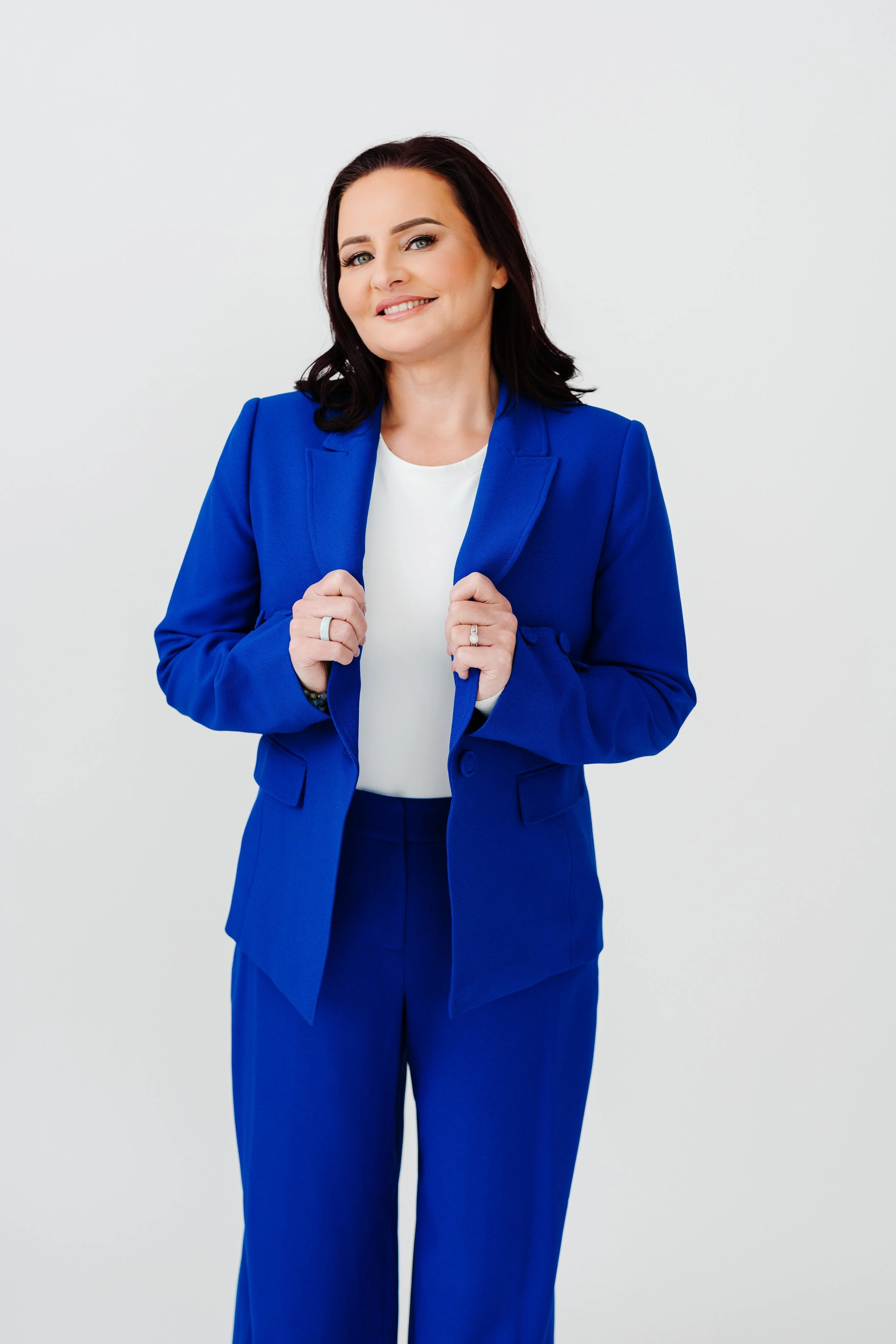 A woman with dark hair, wearing a blue suit and white shirt, smiling and holding the lapels of her blue blazer, standing against a plain light background.