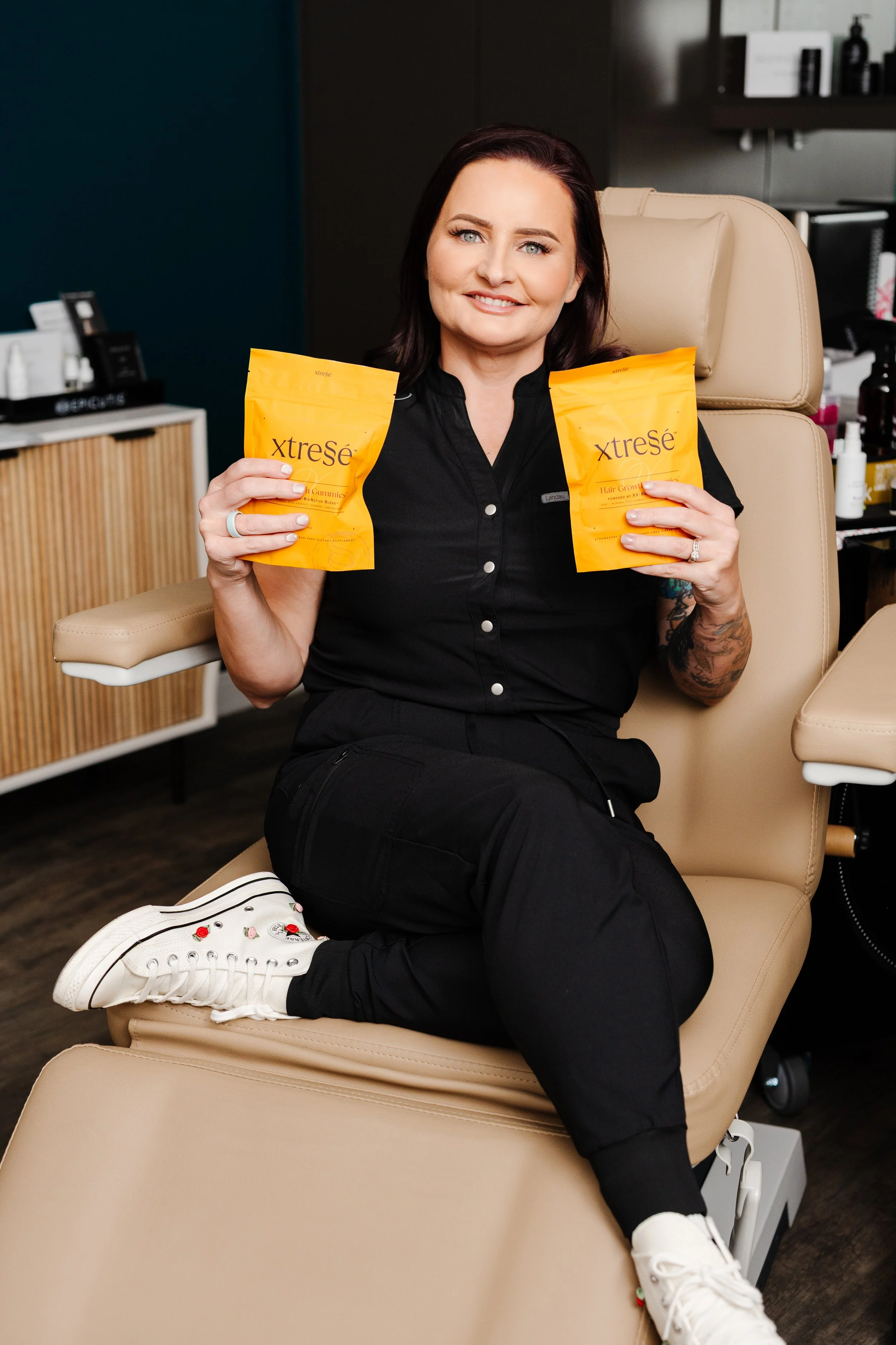 A woman sitting in a beige medical chair, holding two yellow packages labeled 'XtreSé' and smiling at the camera. She is wearing a black shirt, black pants, and white sneakers with cherry and strawberry embellishments.