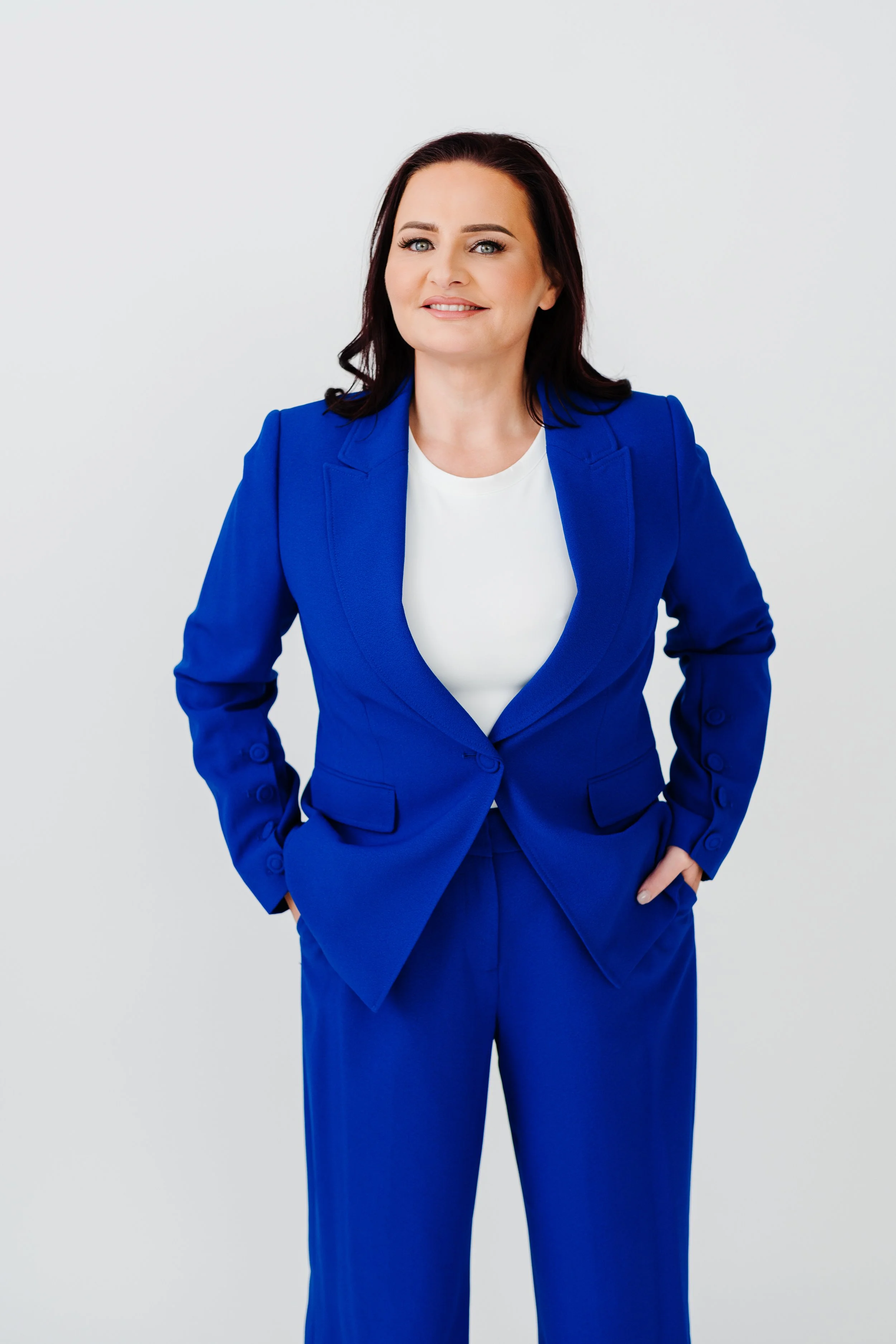 A woman with dark brown hair and blue eyes wearing a bright blue suit and white shirt, smiling confidently, standing against a plain light gray background.