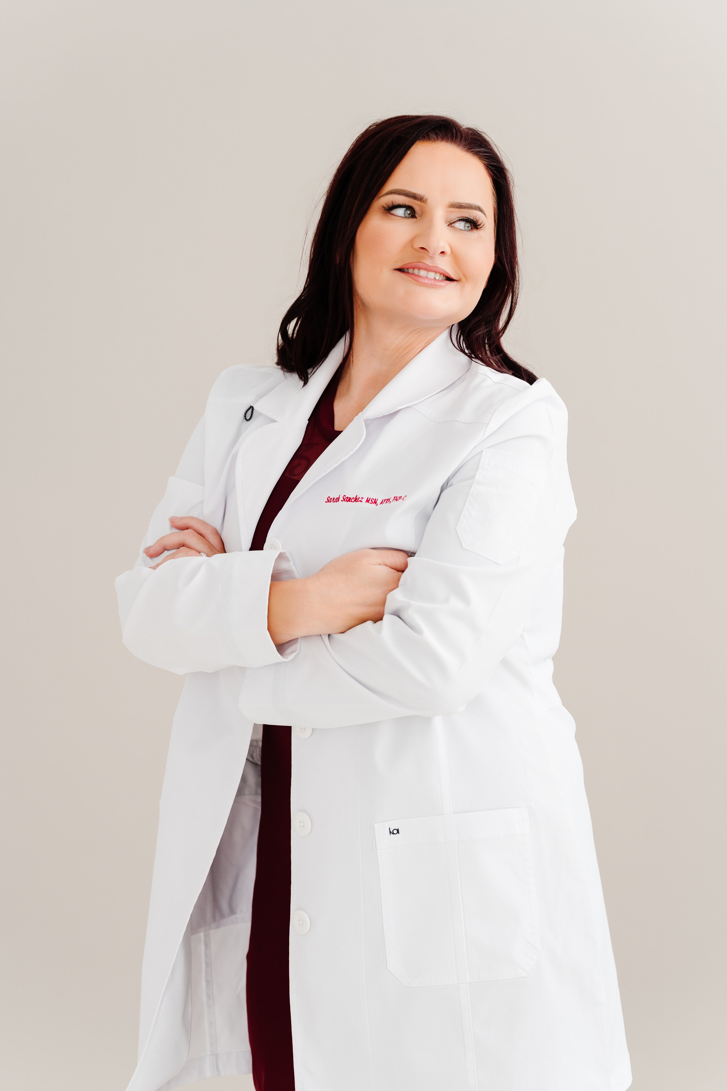 A female doctor with dark brown hair wearing a white lab coat and maroon shirt standing with arms crossed against a plain light beige background.
