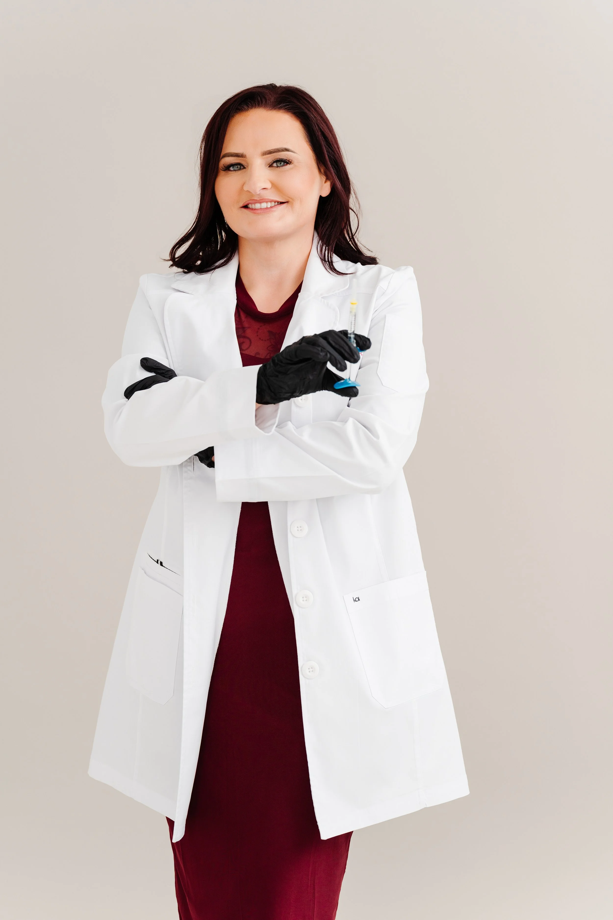 A woman with shoulder-length dark red hair, wearing a white lab coat over a burgundy dress, holding a syringe, smiling against a plain background.