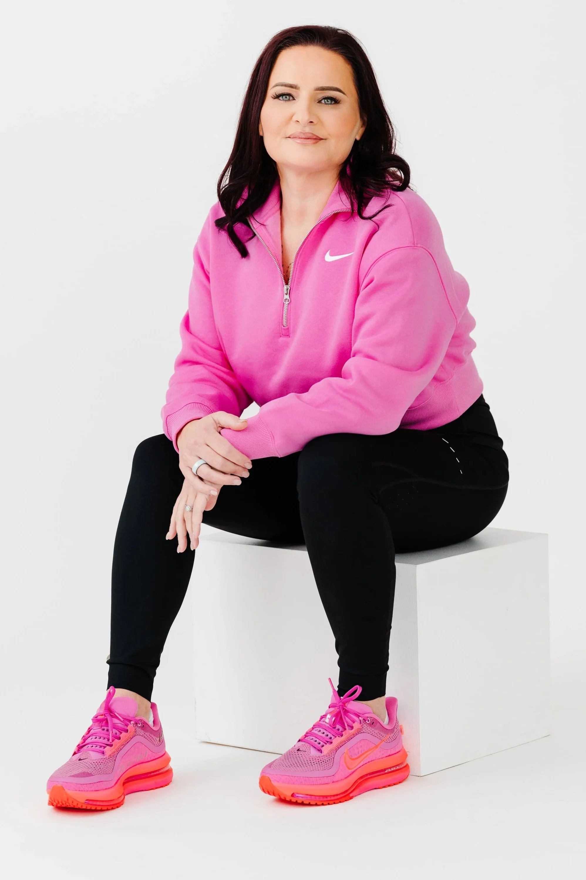 A woman wearing a pink Nike zip-up hoodie, black pants, and bright pink sneakers sitting on a white block against a plain white background.