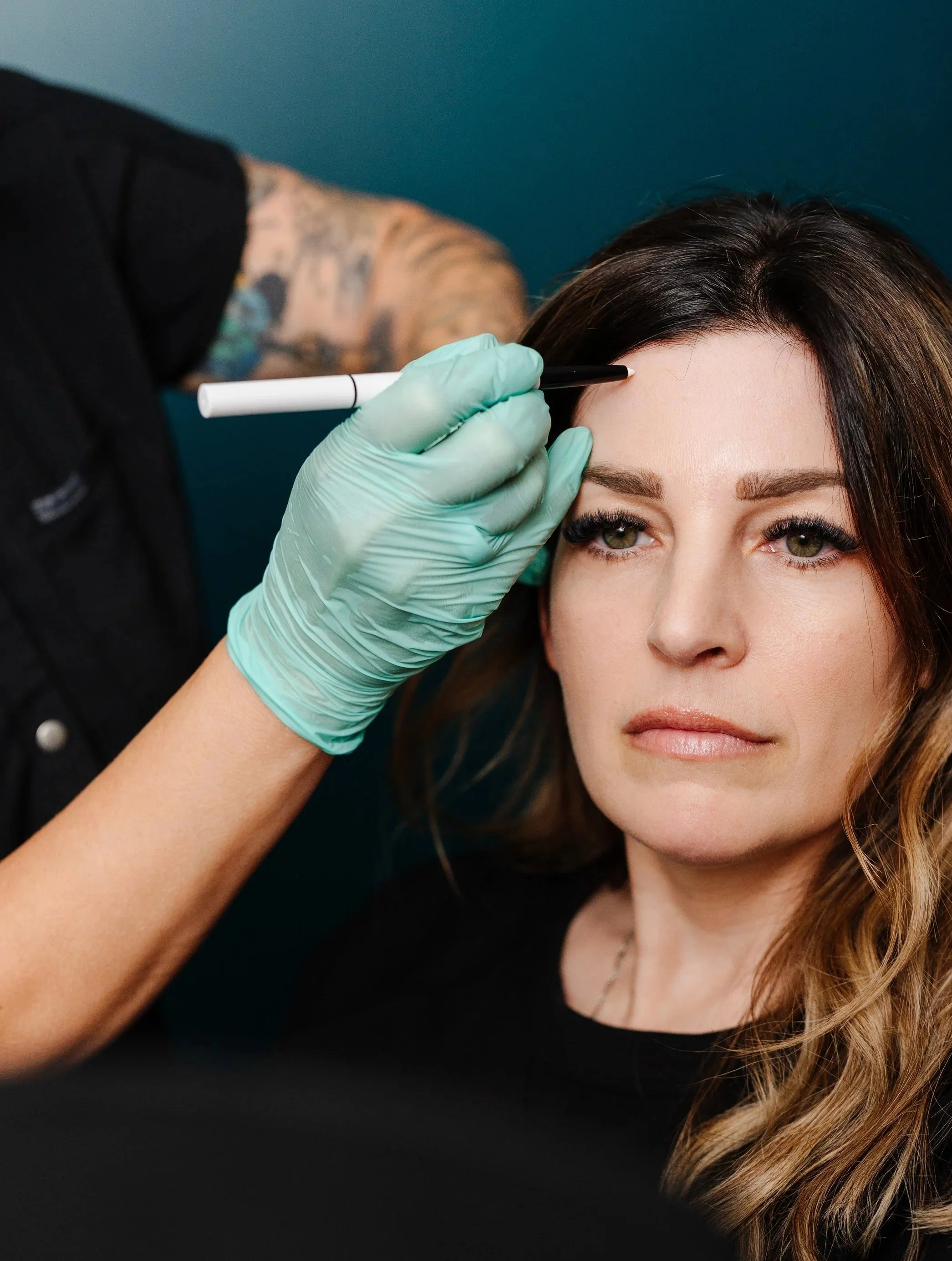 A woman with curly, light brown hair and green eyes having her eyebrows tattooed by a tattoo artist using a tattoo pen. The tattoo artist is wearing a black shirt and green gloves.