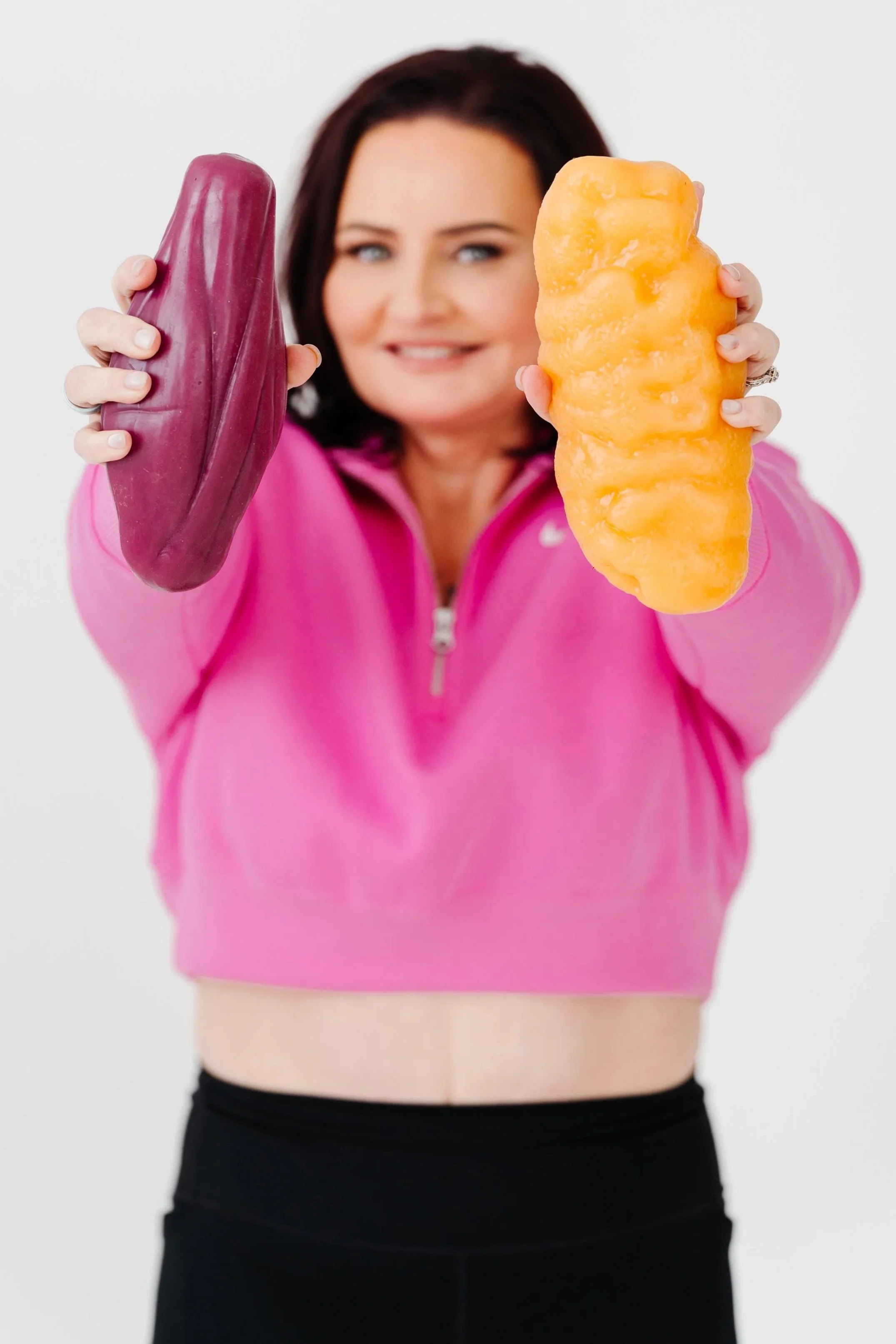 Woman with short dark hair wearing a pink zip-up top, holding two large pieces of sweet potato or squash in front of her, one purple and the other orange, against a plain white background.