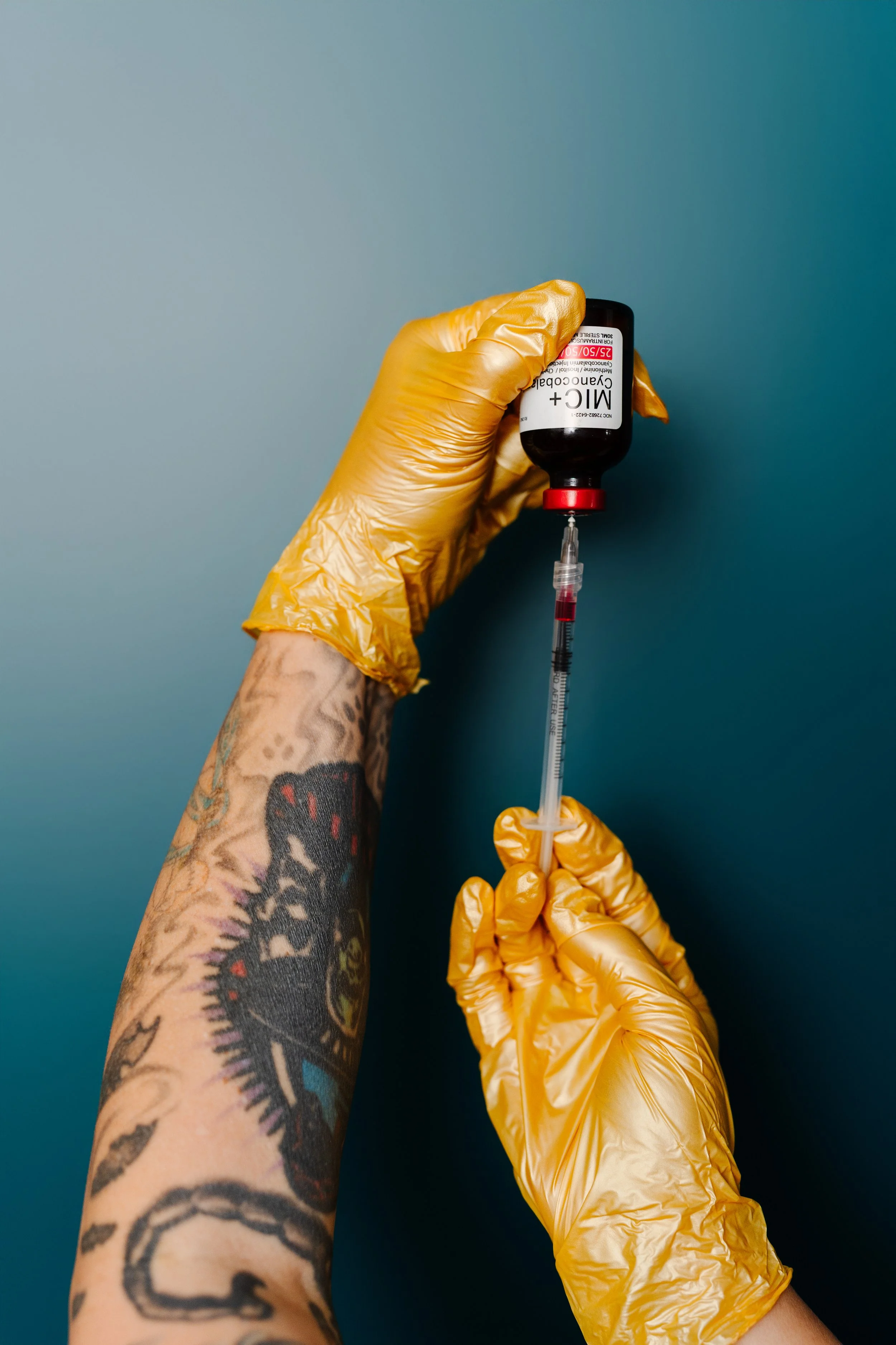 A tattooed arm wearing yellow gloves holding a medical vial with a syringe attached against a teal background.