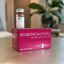 A vial of Sculptra Aesthetic resting on a pink box labeled 'Sculptra Aesthetic injectable poly-L-lactic acid,' placed on a wooden surface with a blurred background of a plant and room interior.