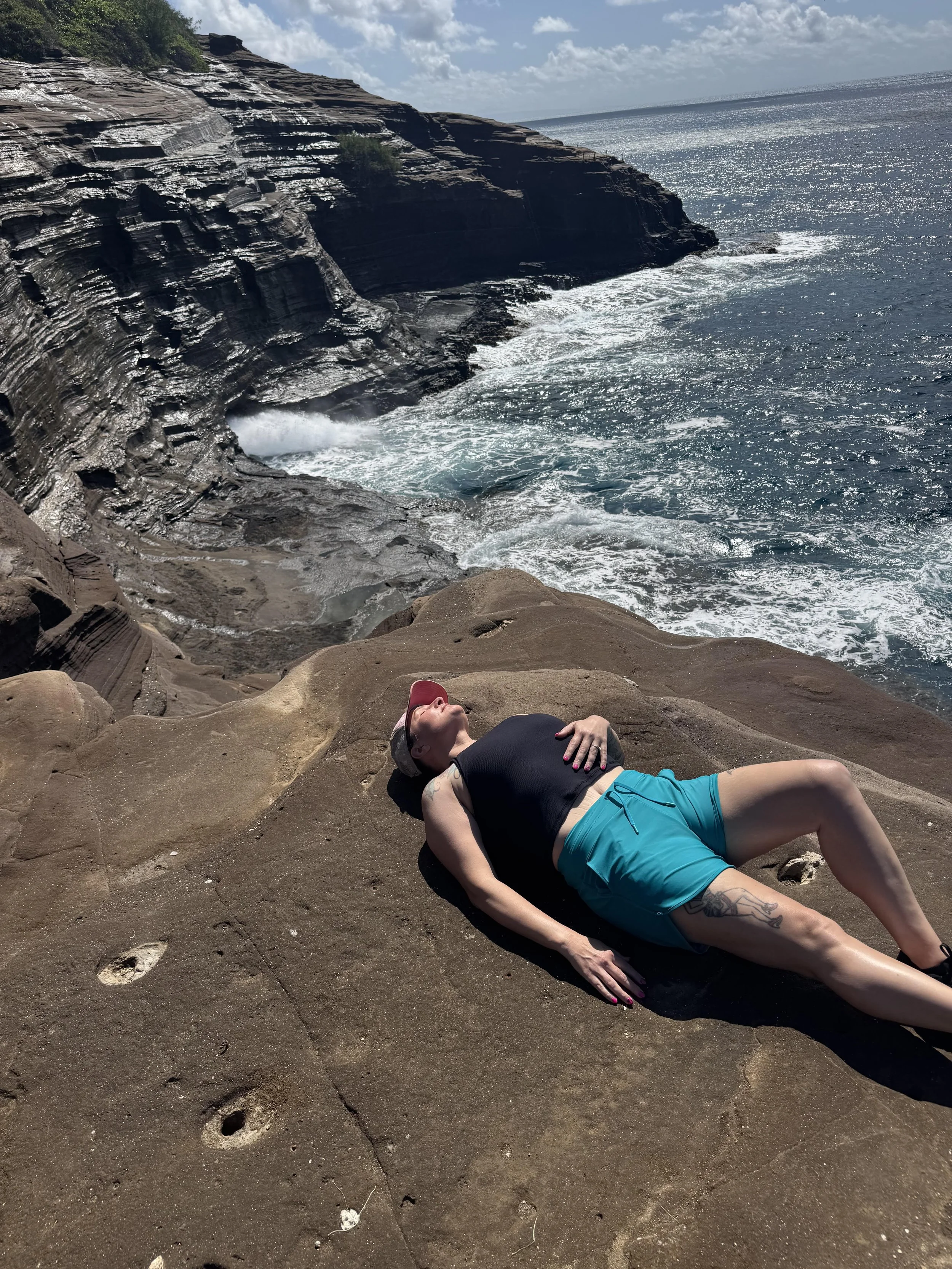 A woman lying on a rocky beach with ocean waves crashing nearby. She is wearing a black tank top, blue shorts, a pink cap, and sunglasses, with her hands resting on her chest and her head tilted back.