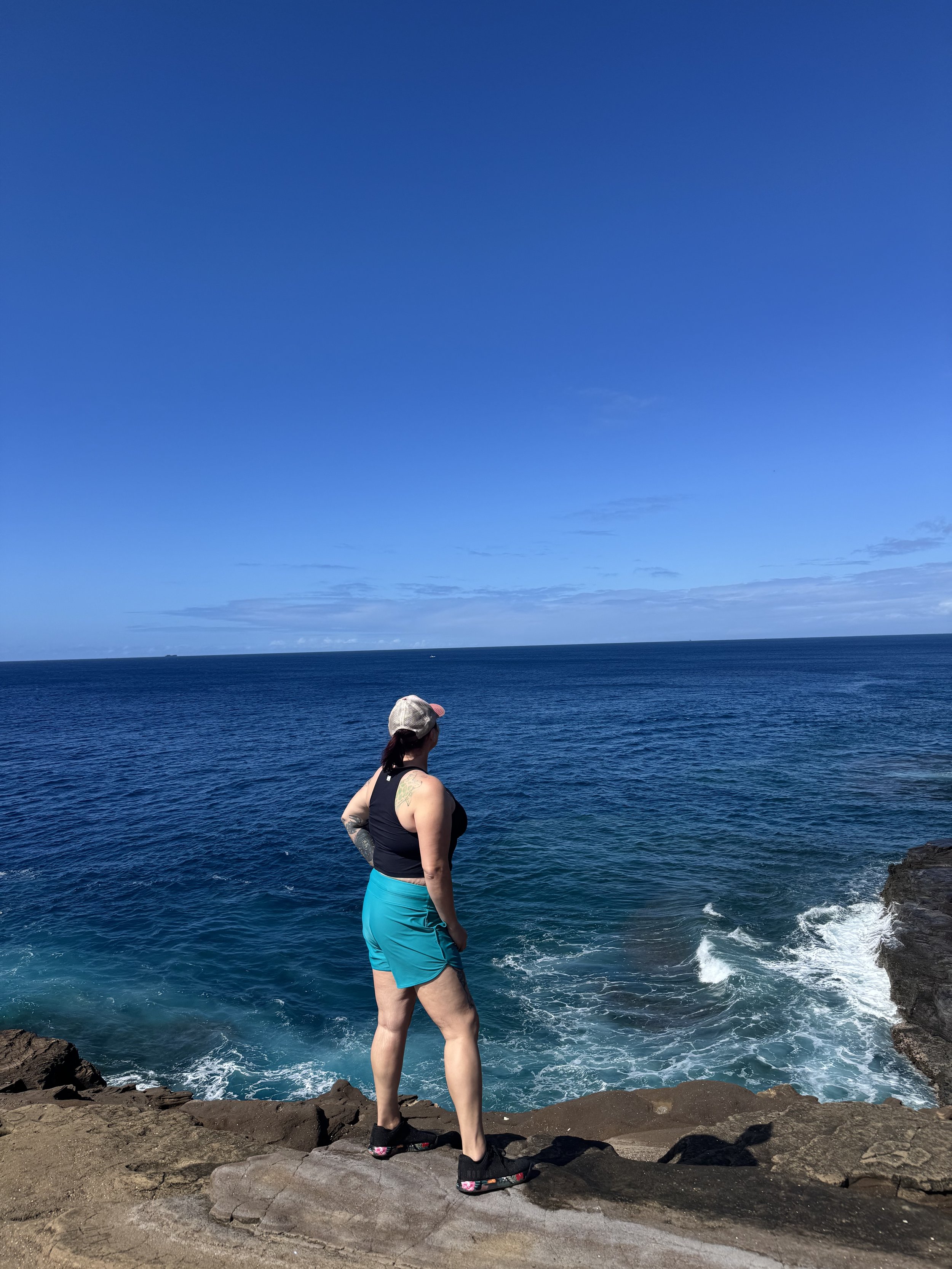 A woman standing on rocky terrain near the ocean, looking out over the water and blue sky.