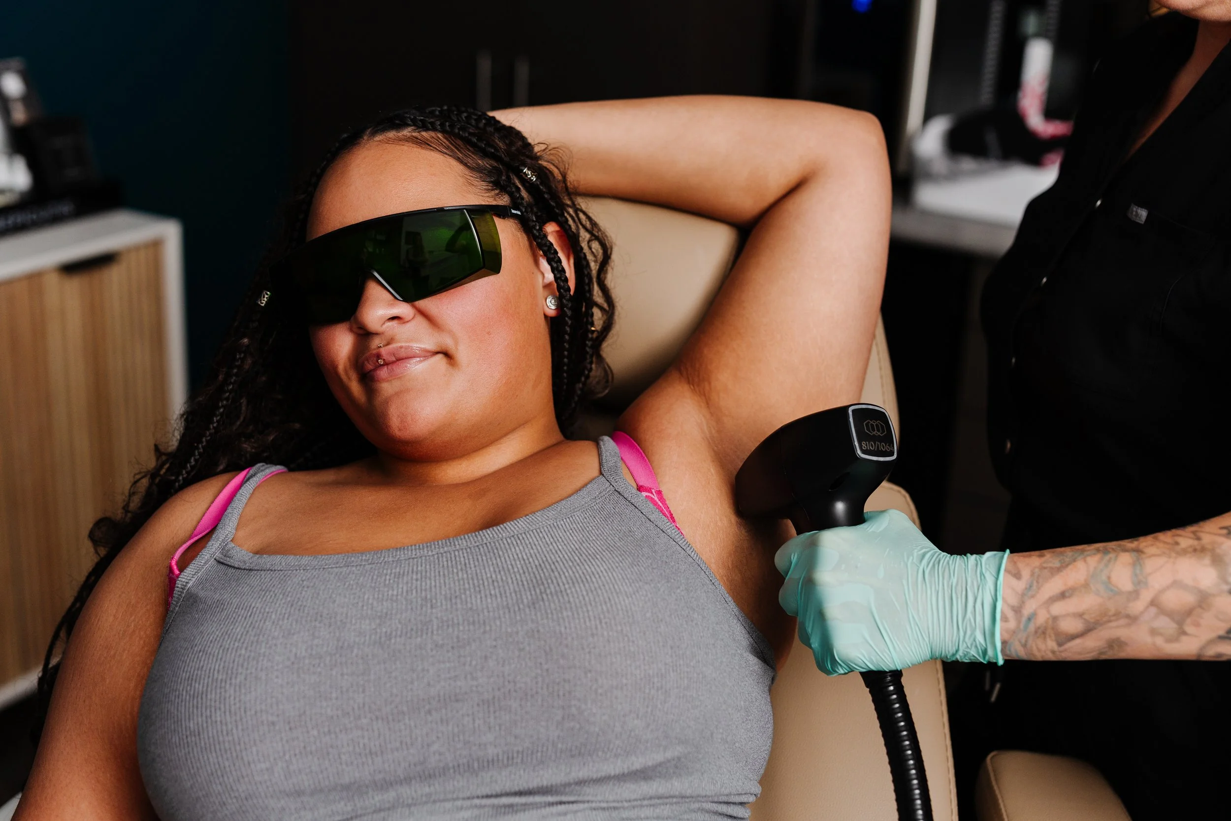 A woman with braided hair and sunglasses lying back while receiving a medical injection in her upper arm, with a healthcare professional administering the shot using a syringe and wearing gloves.