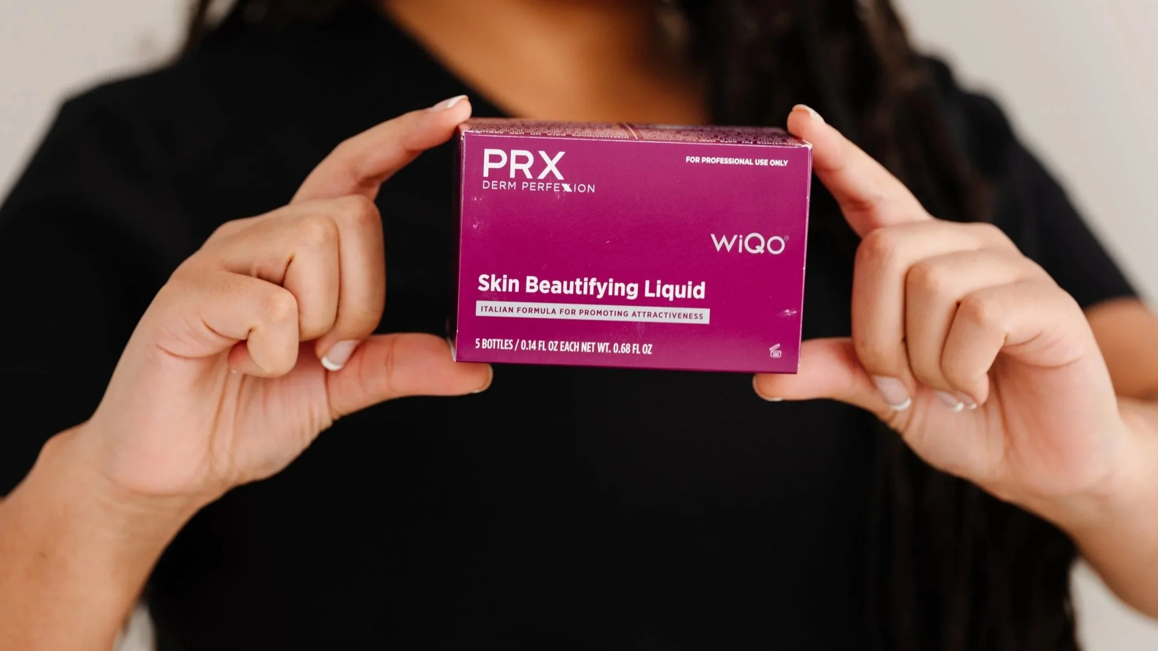 A person holding a purple box labeled 'PRX Derm Perfection WiQo Skin Beautifying Liquid' with text indicating it is for professional use only and contains five small bottles.