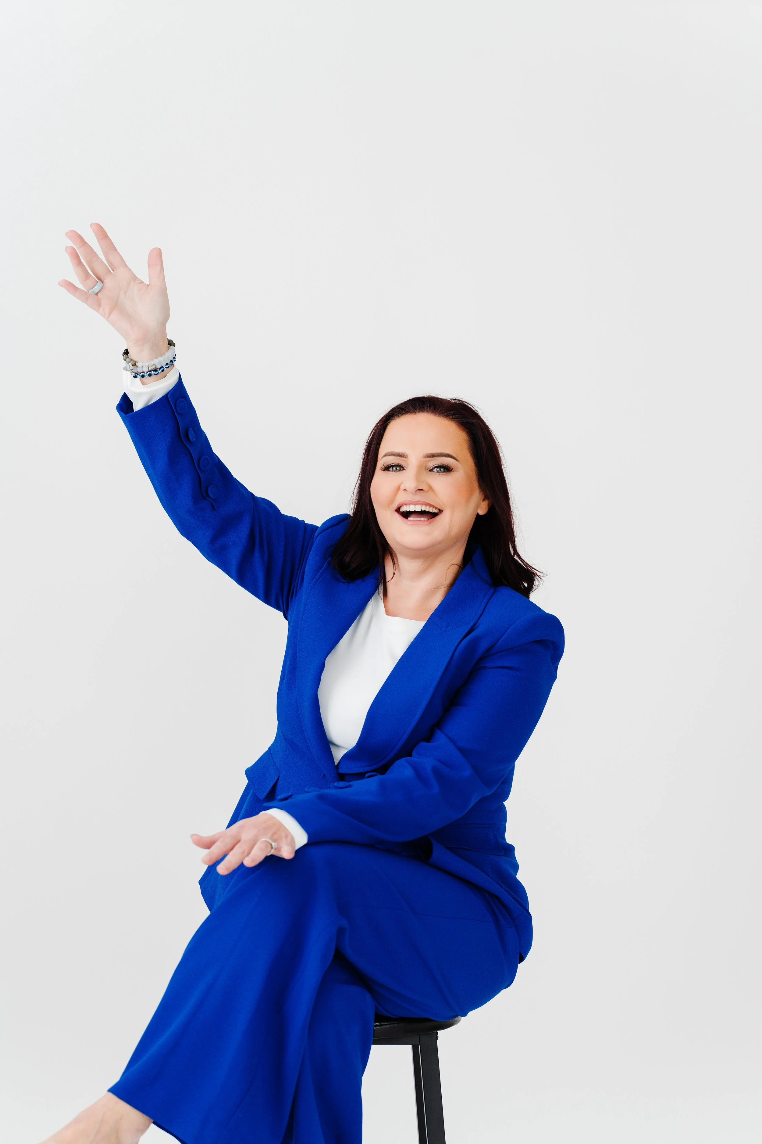 A woman with dark brown hair wearing a bright blue suit, sitting on a black stool, smiling and waving with her right hand, against a plain white background.