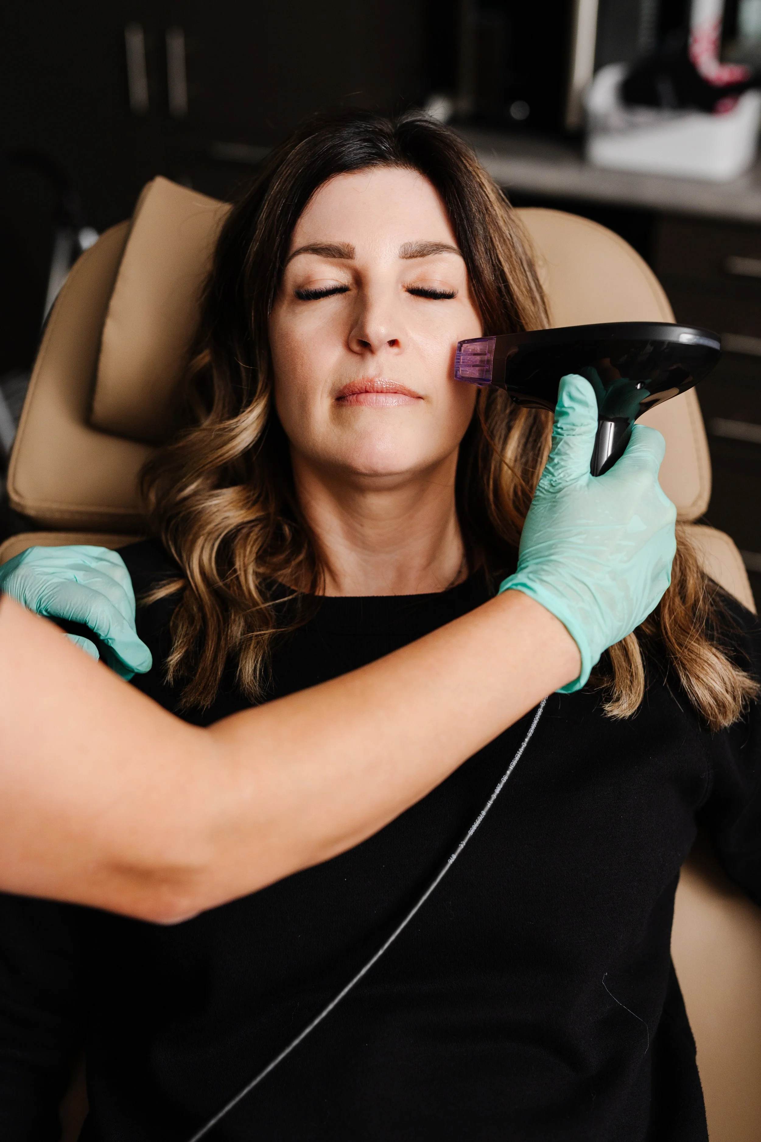 A woman with long wavy brown hair and closed eyes is lying on a beige massage chair with her head resting on a pillow. She is receiving facial treatment or skincare therapy from a professional wearing green gloves who is holding a small handheld device near her face.