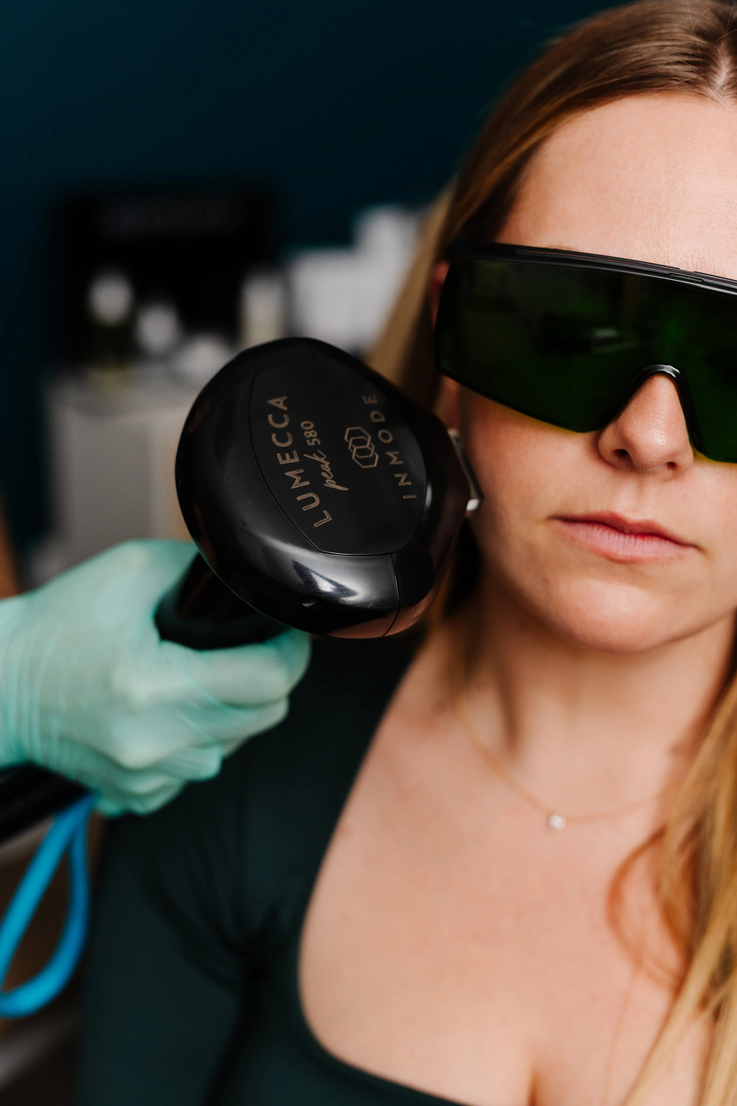 A woman wearing sunglasses and a black top receiving a skin consultation with a handheld device by a person wearing gloves.