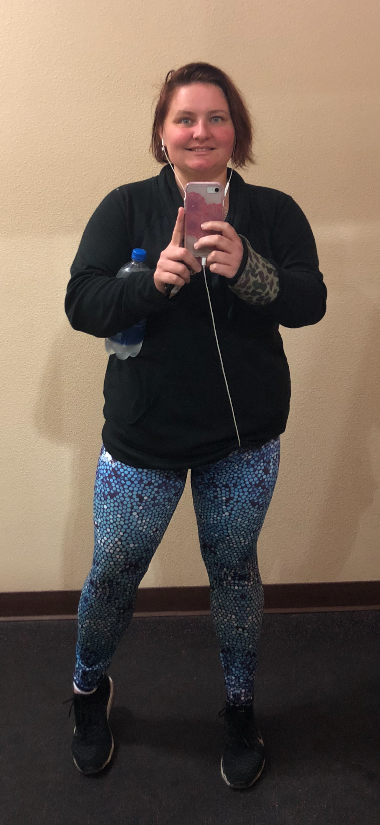 Person taking a selfie in workout clothes, holding a water bottle and phone, wearing earbuds, with a neutral background.
