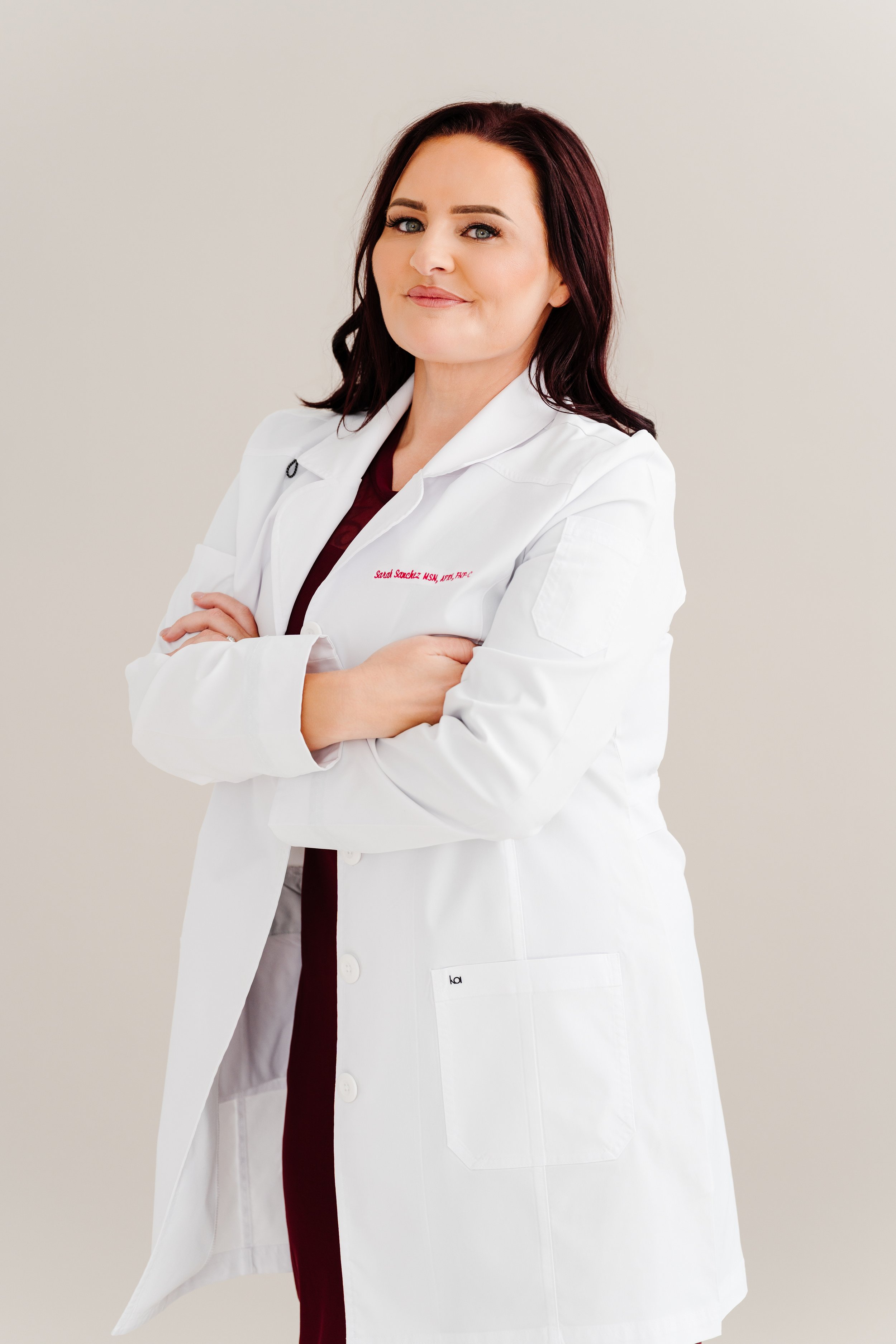 A female doctor standing with arms crossed, wearing a white lab coat over maroon scrubs, smiling confidently against a plain white background.