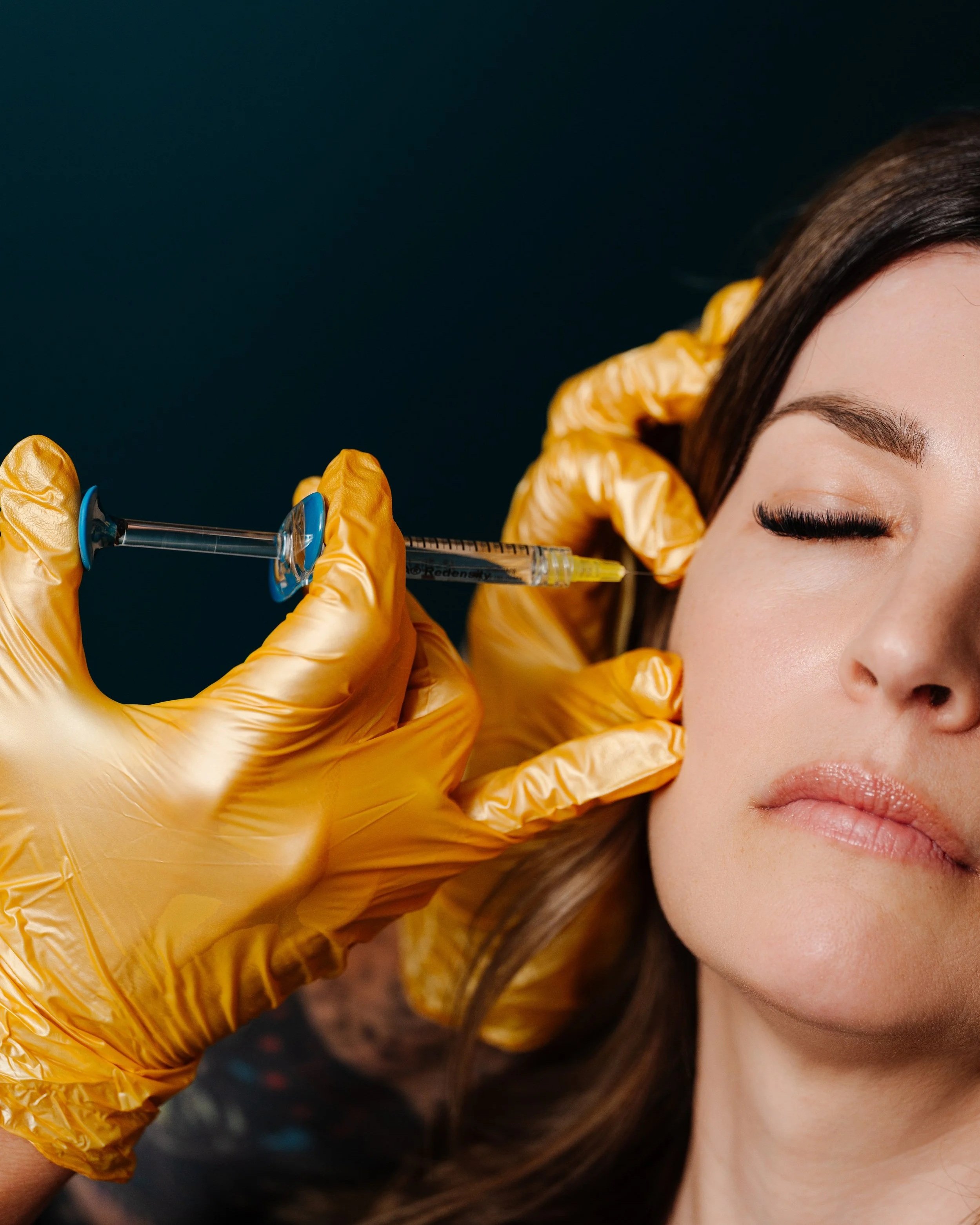 Close-up of a woman with closed eyes receiving a cosmetic injection in her face as a medical professional wearing yellow gloves holds the syringe.