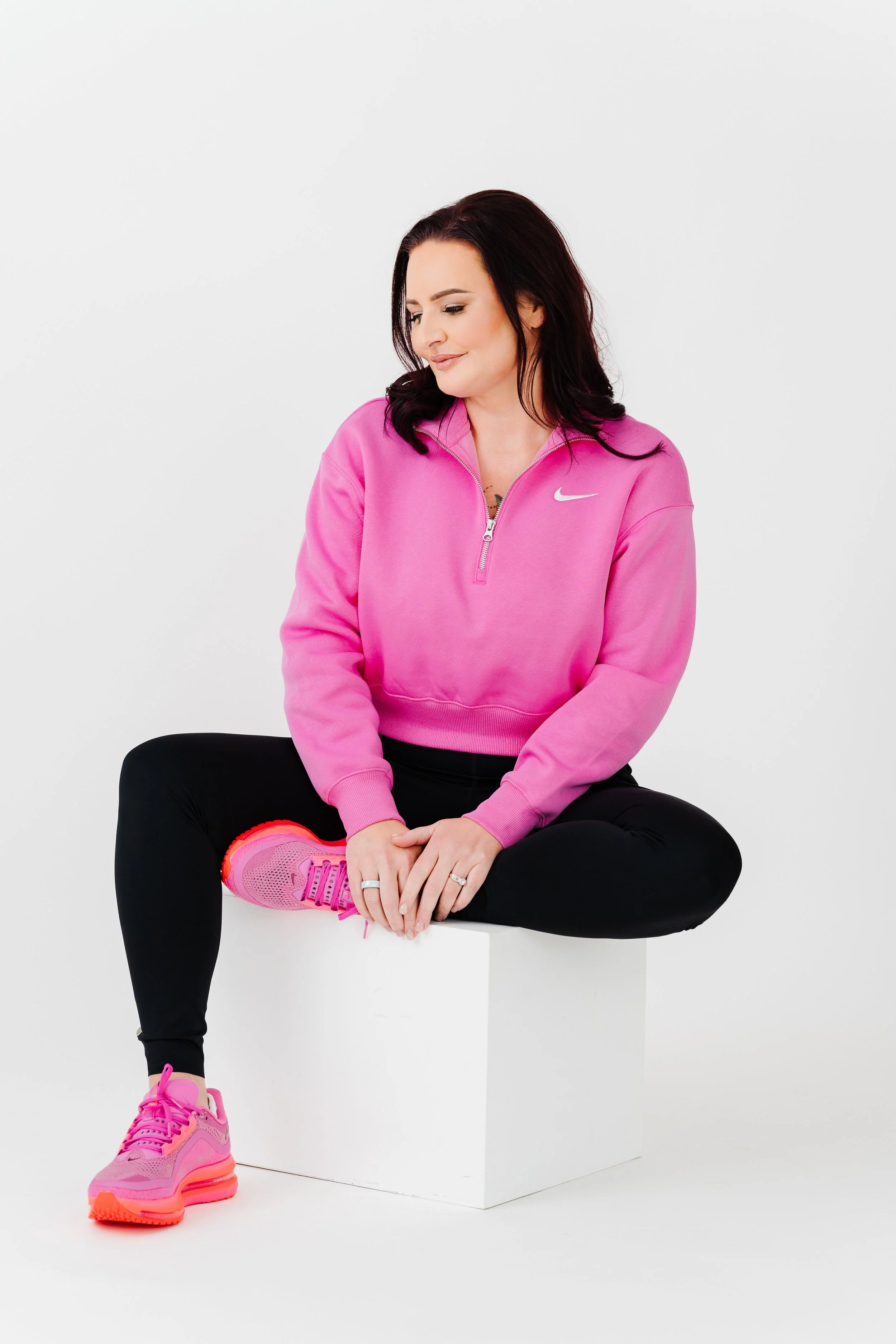 Woman dressed in pink Nike athletic top and black pants sitting on a white block with one leg crossed over her lap, wearing bright pink sneakers, against a plain white background.