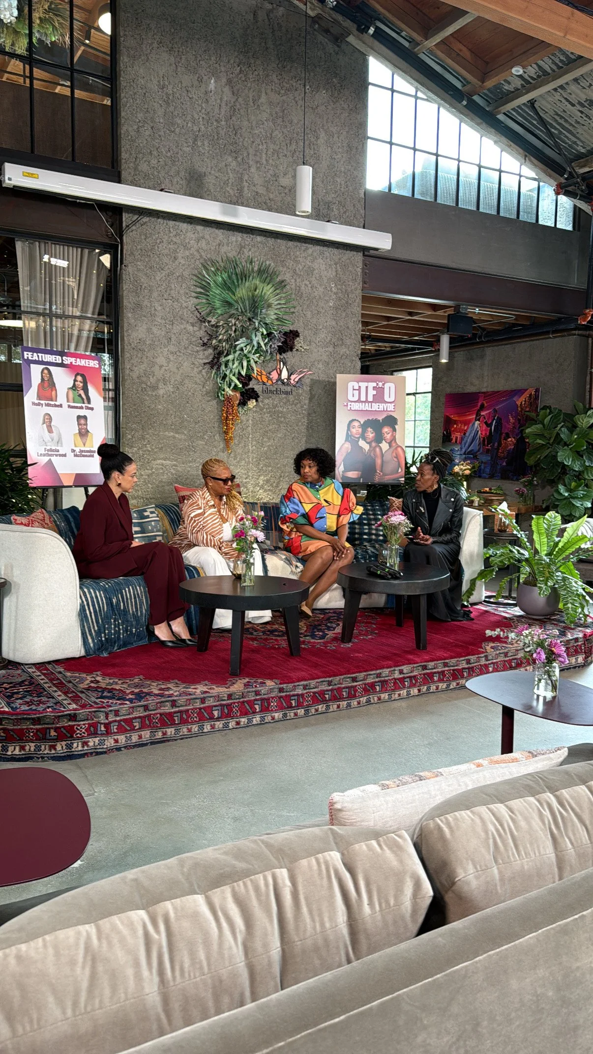 Four women sitting on a couch in a stylish, modern indoor space, engaging in conversation. The space features contemporary decor, large windows, potted plants, and promotional posters on the wall behind them.