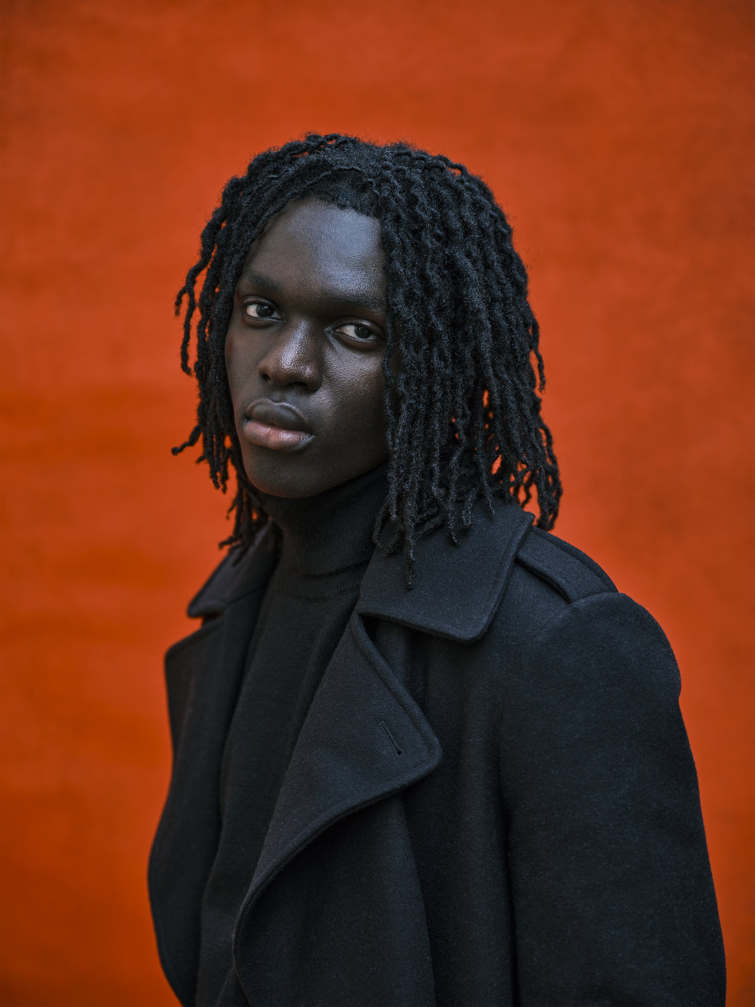 Portrait of a black woman with shoulder-length dreadlocks, wearing a black coat and turtleneck, against an orange background.