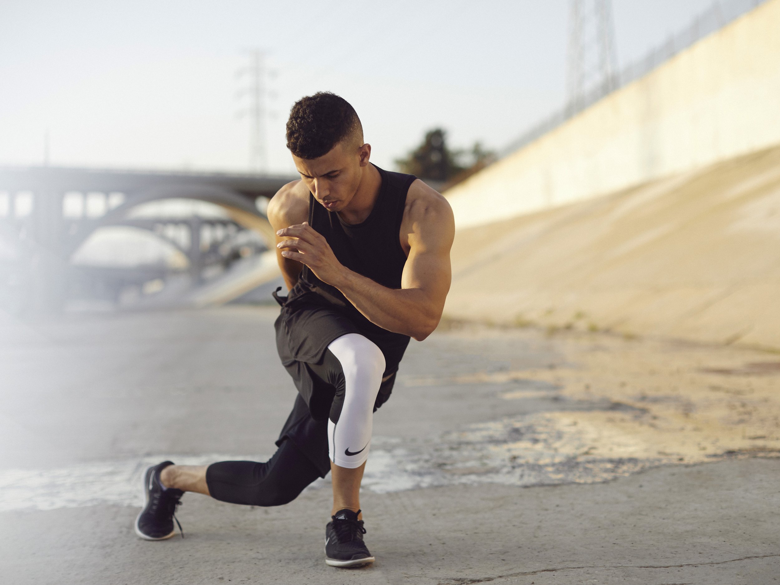 A young man stretching outdoors on a paved surface near a bridge and a wall, dressed in athletic gear, preparing for exercise.