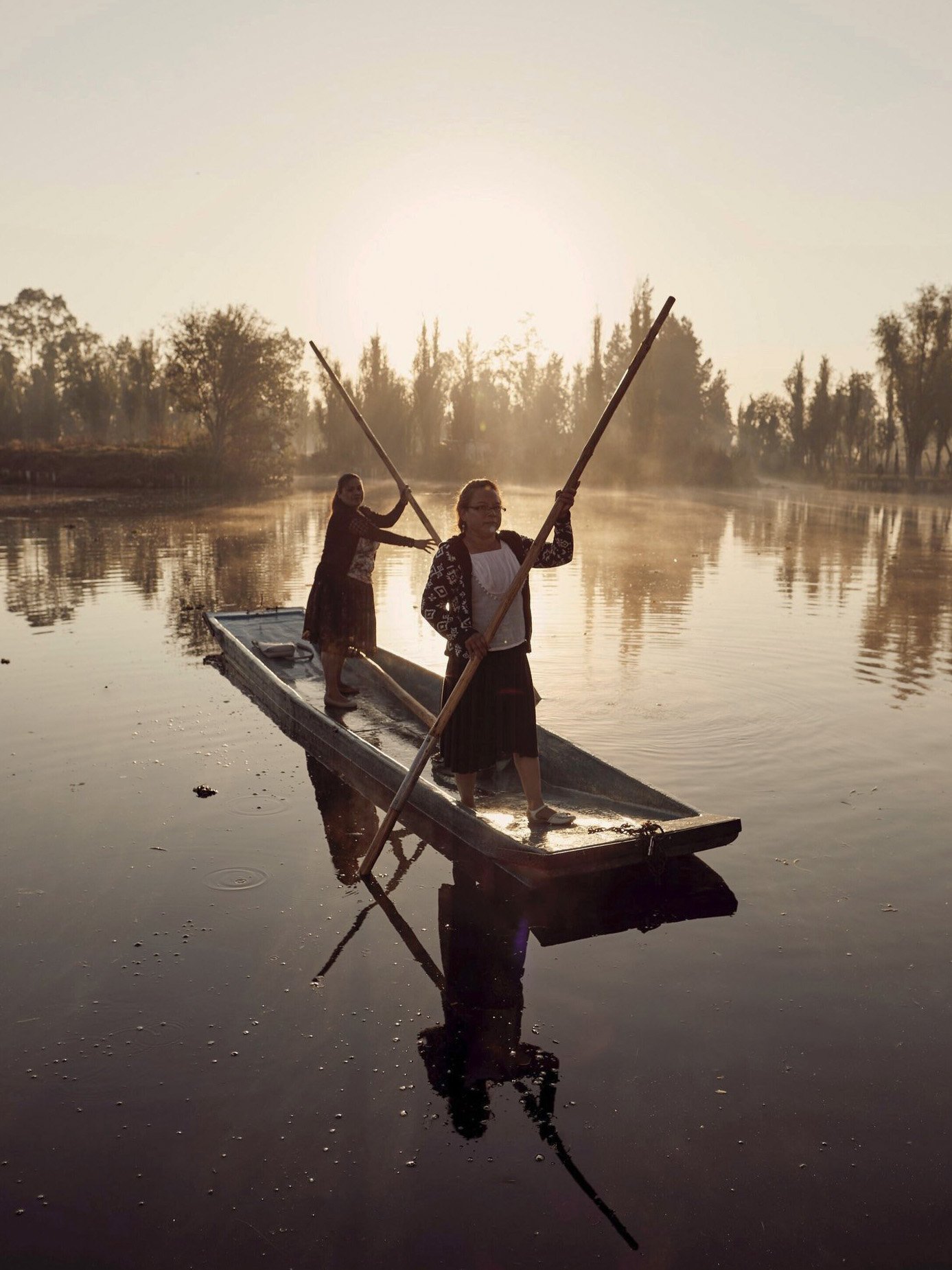 Two women standing on a boat on a calm river at sunset, using long poles to navigate, with trees in the background and their reflection visible in the water.