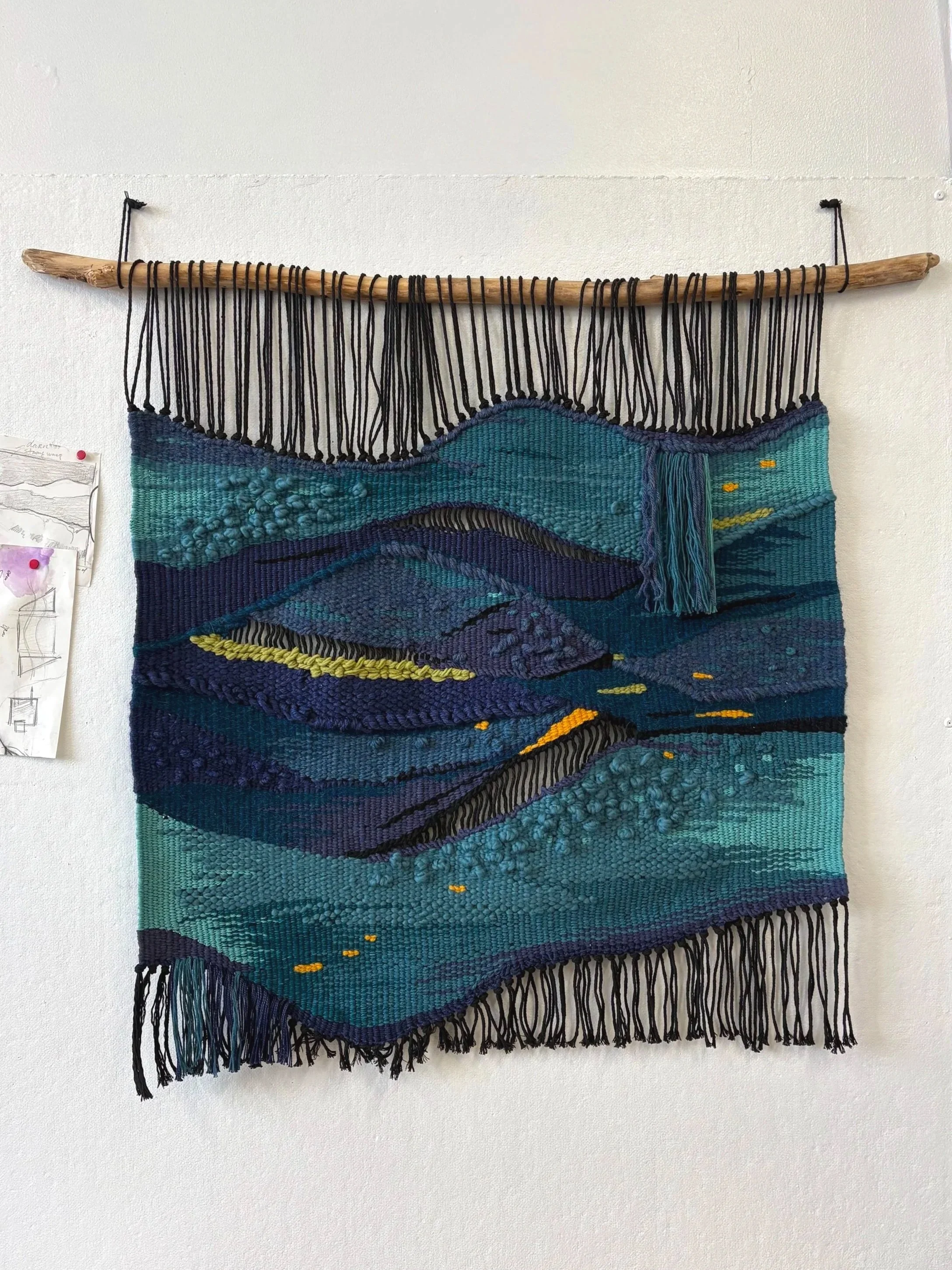 Wall hanging textile art depicting a landscape with mountains, water, and sky in shades of blue, teal, purple, and yellow, suspended from a wooden dowel with black hanging cords.