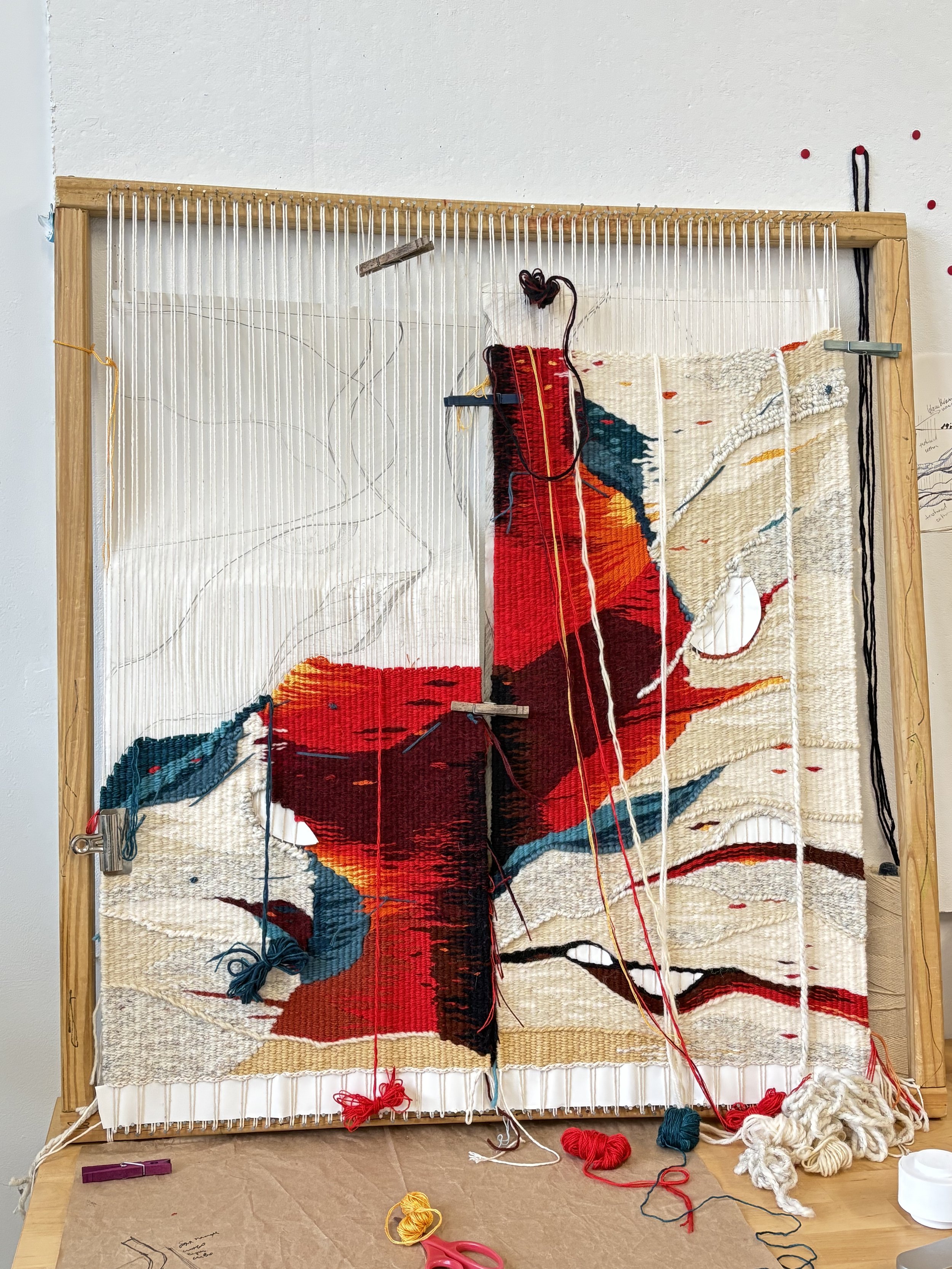 A textile art piece in progress mounted on a wooden frame, with colorful yarns, knitting needles, scissors, and a partially finished woven tapestry featuring a stylized red and orange bird or creature, on a beige background.