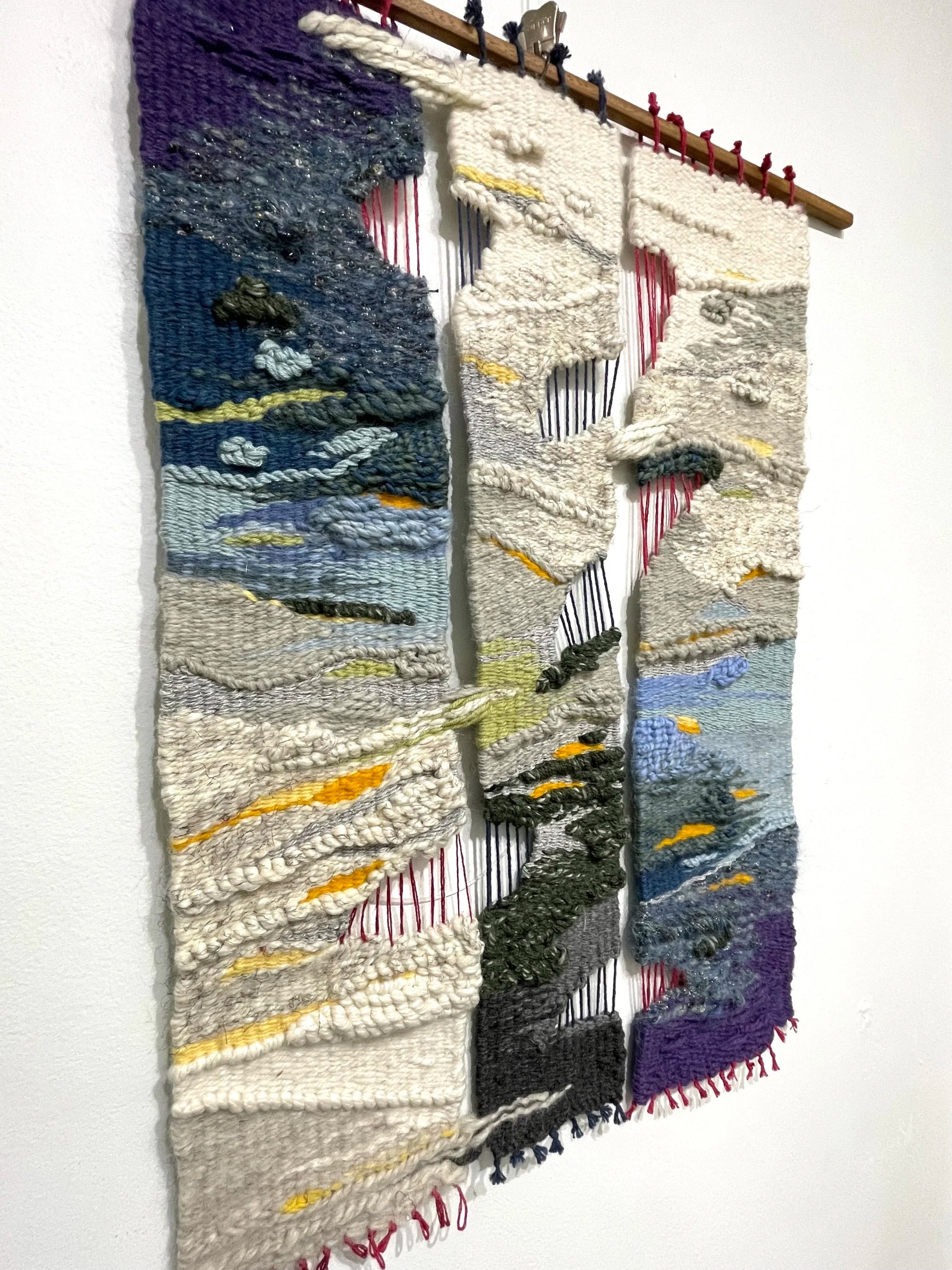 A textured woven tapestry artwork depicting abstract landscape with clouds, mountains, and sky in shades of blue, gray, beige, yellow, purple, and black, hanging on a wall.