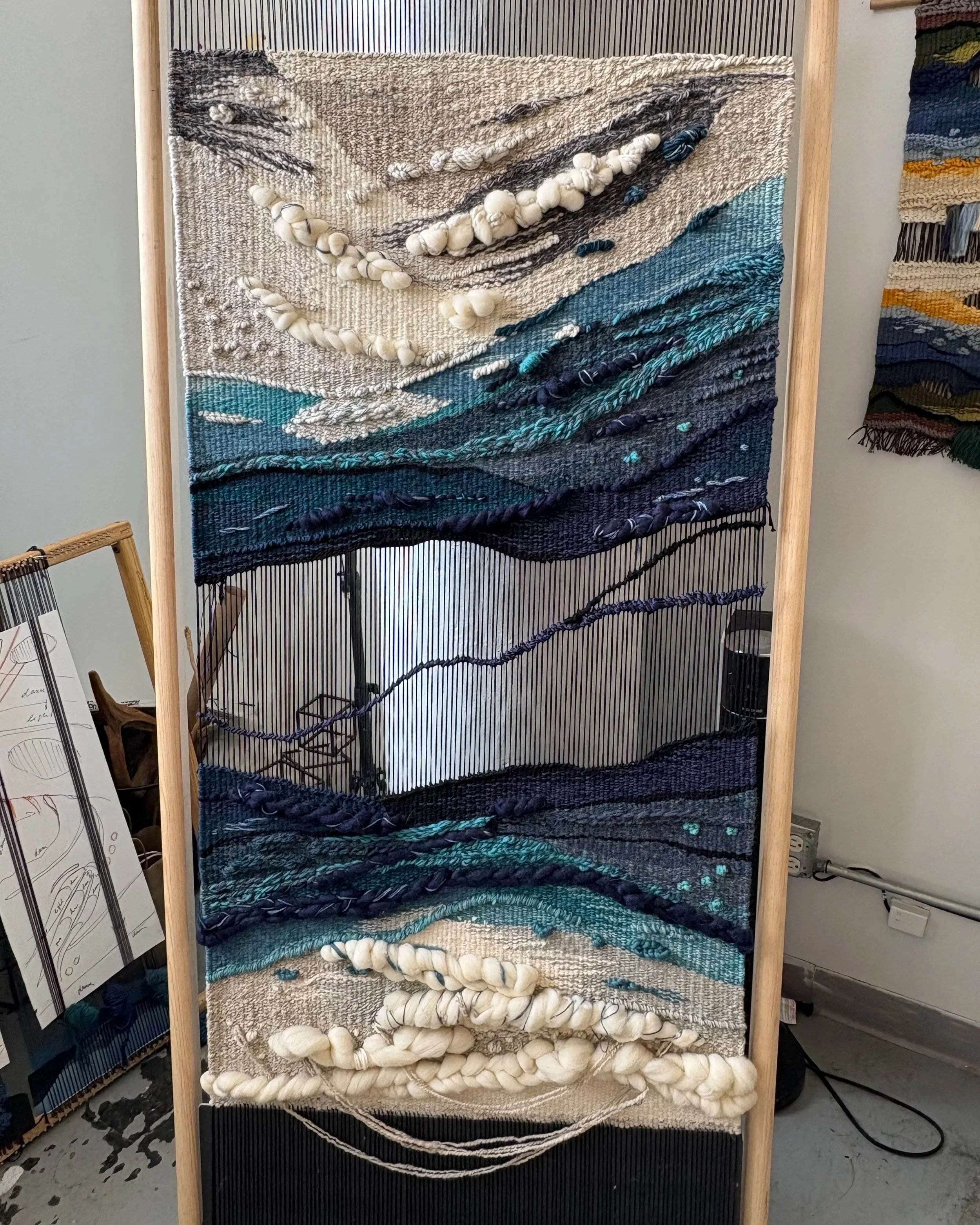 A handmade woven wall hanging art piece with abstract landscape design, mounted on a wooden frame, featuring textured yarn and vibrant blue, turquoise, black, and cream colors.
