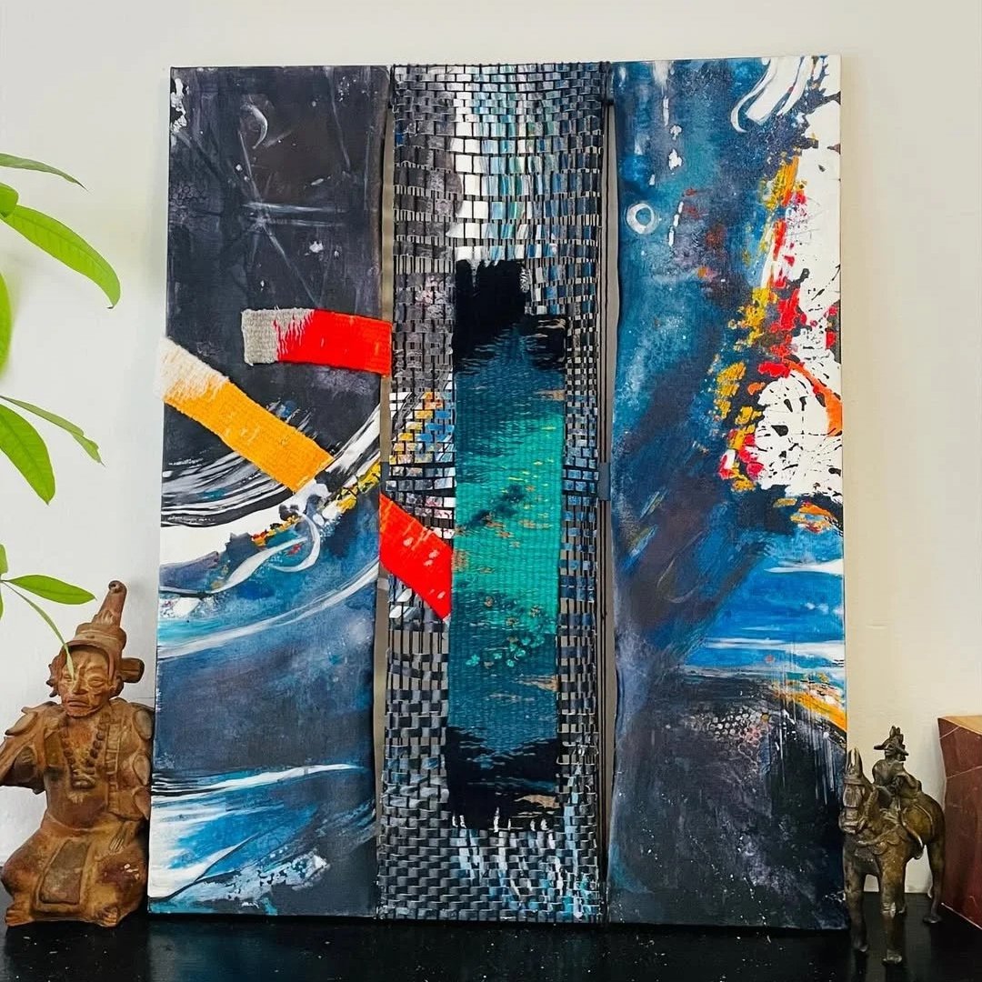 An abstract mixed media artwork with vibrant colors, textured patterns, and three-dimensional elements, displayed on a wall with sculptures and a plant nearby.