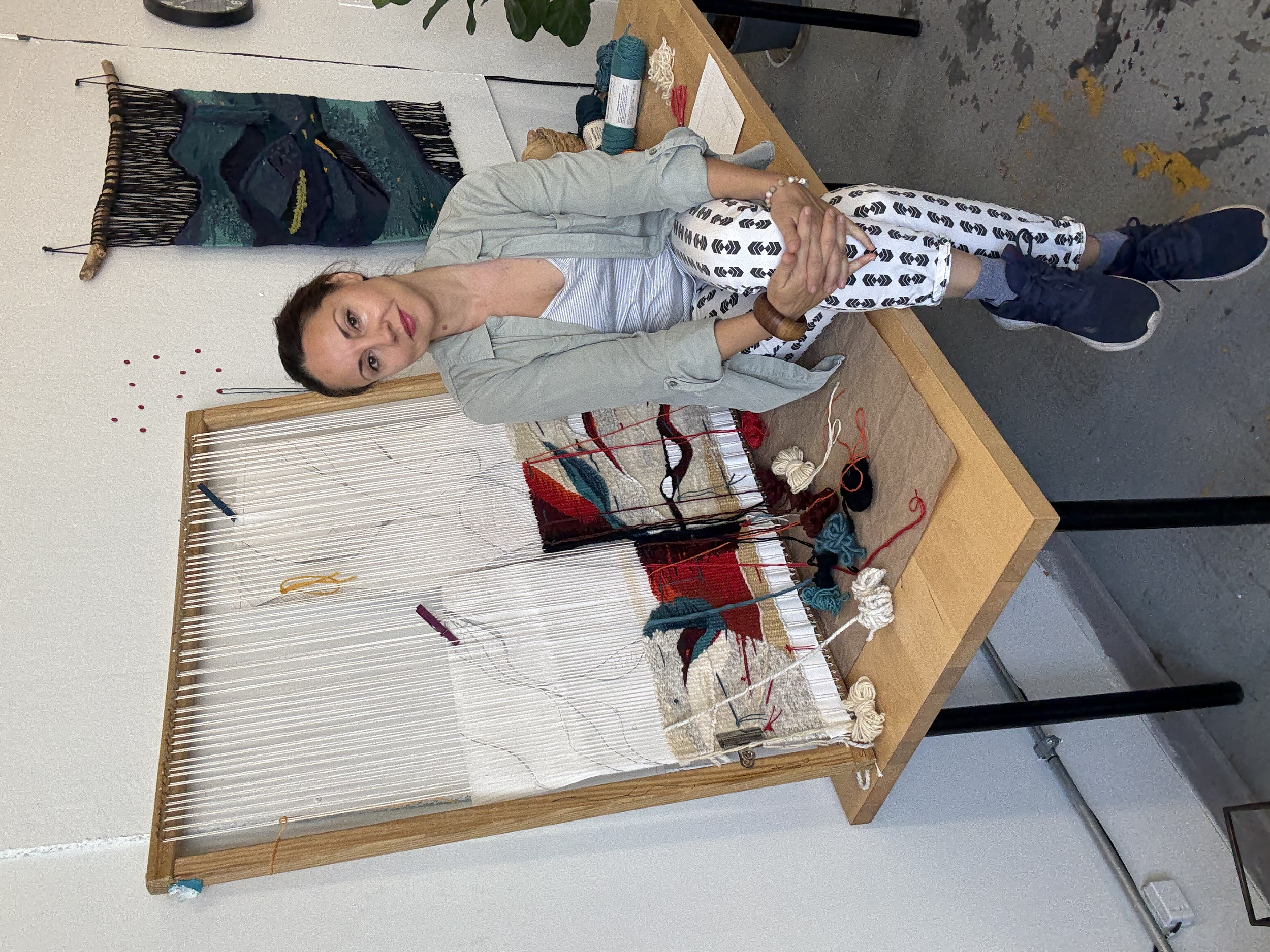 A woman sitting at a table with a weaving loom in front of her, surrounded by balls of yarn in various colors.
