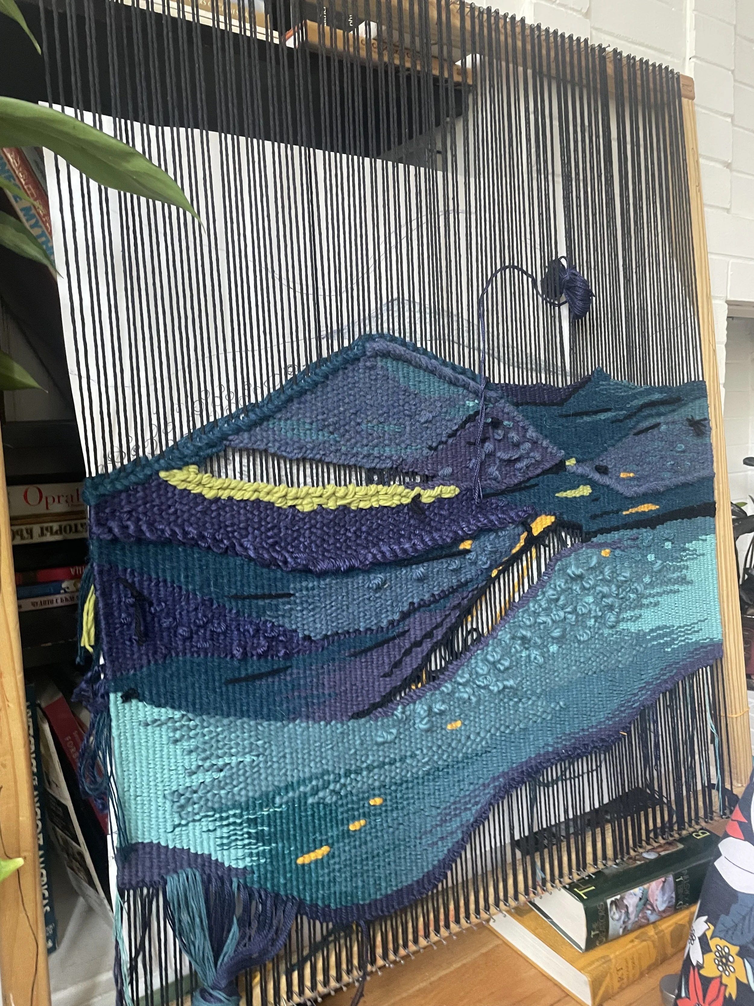 A large, colorful woven tapestry in progress on a loom. The design features shades of blue, purple, and yellow, resembling a landscape or abstract scene.