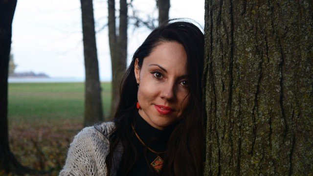 Woman with dark hair and red lipstick leaning against a tree outdoors in a wooded area near a body of water.