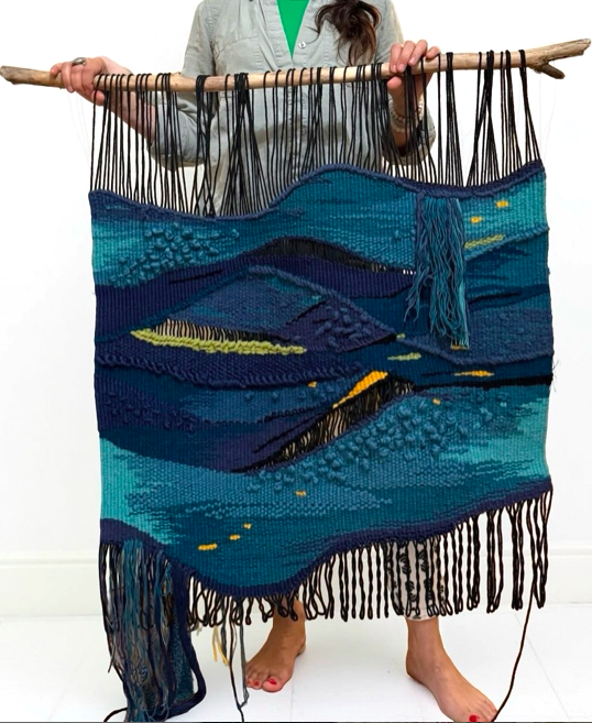 Person holding a large, woven landscape tapestry with a wooden rod, depicting a night scene with mountains, water, and stars.
