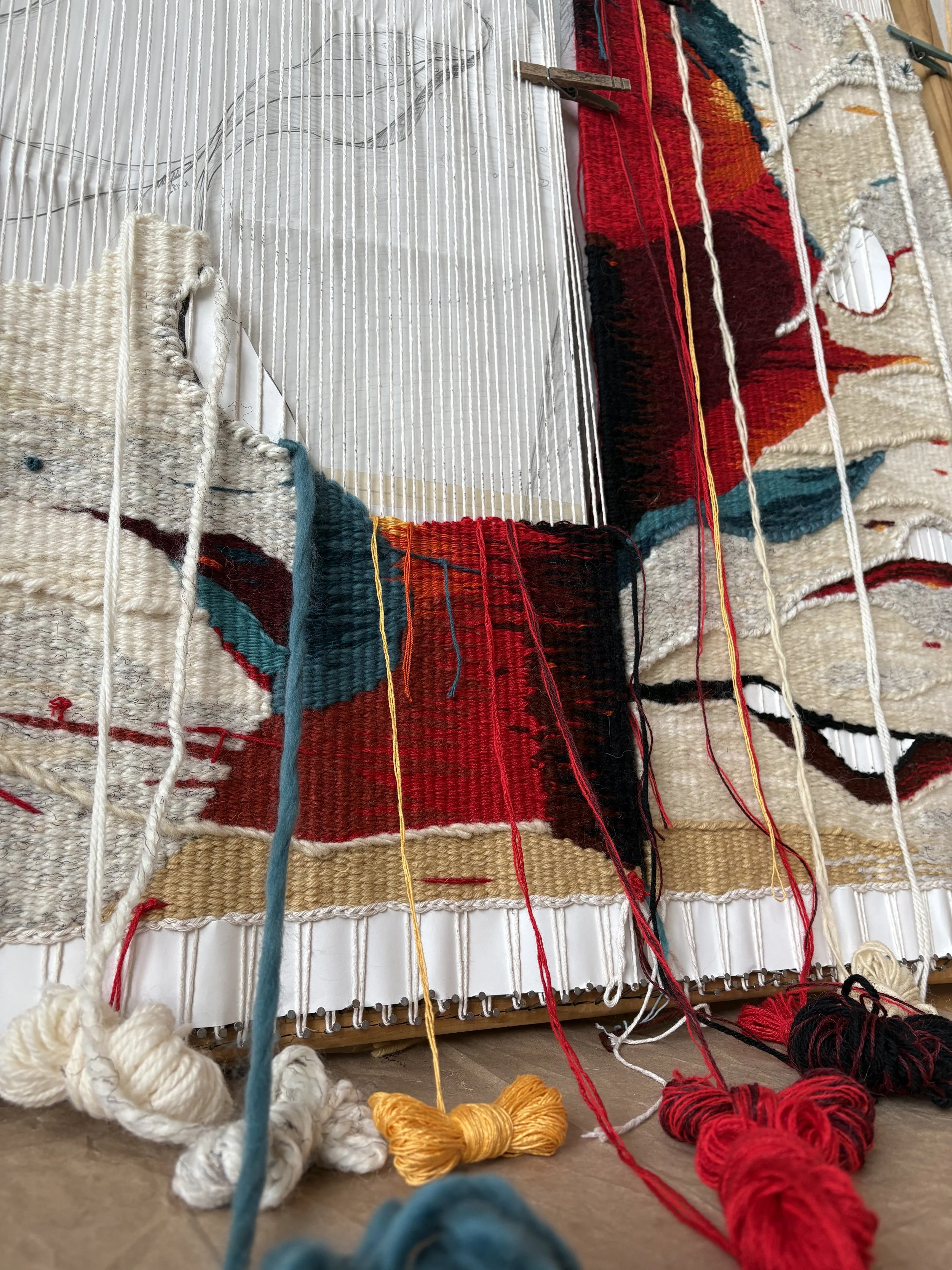 Close-up of a weaving loom with partially woven colorful textiles, including red, black, and cream patterns, and yarns of various colors hanging from the loom.