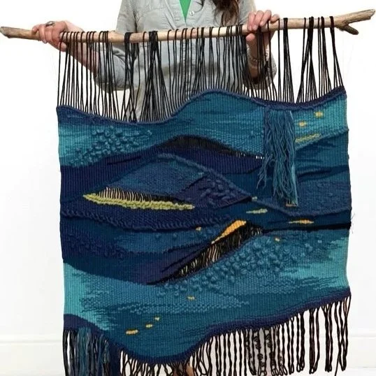 A person holding a large handwoven tapestry or wall hanging with a landscape scene in shades of blue, green, and yellow, attached to a wooden rod.